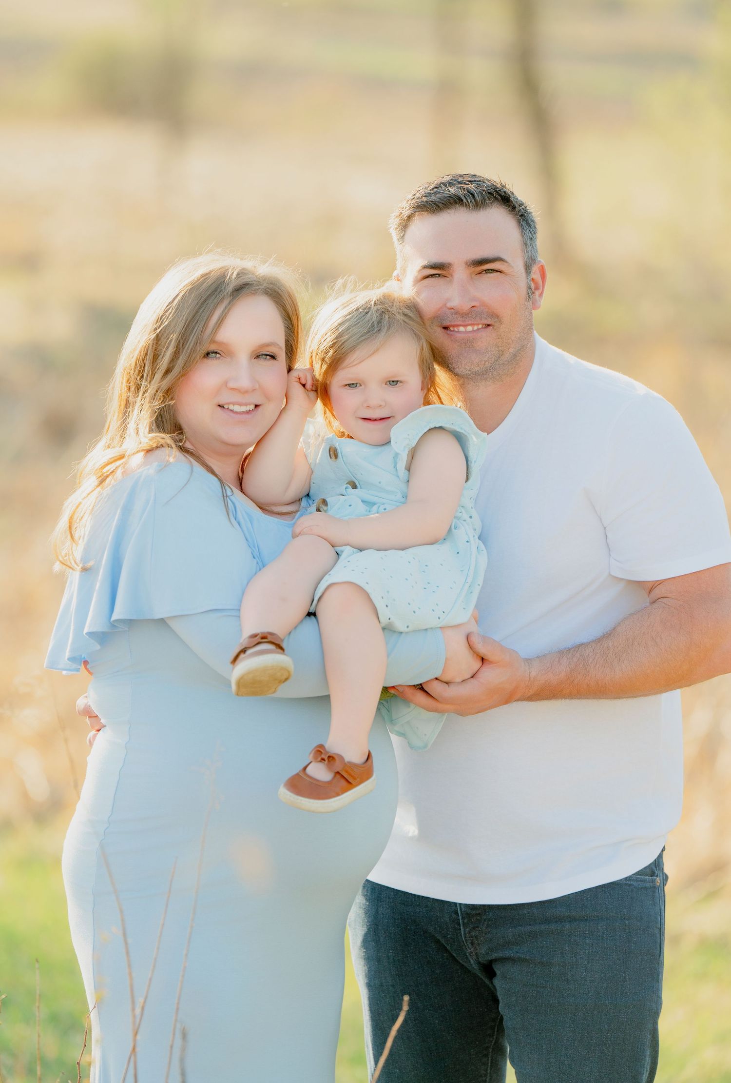 A family of three poses outdoors during golden hour in coordinating blue and white outfits.