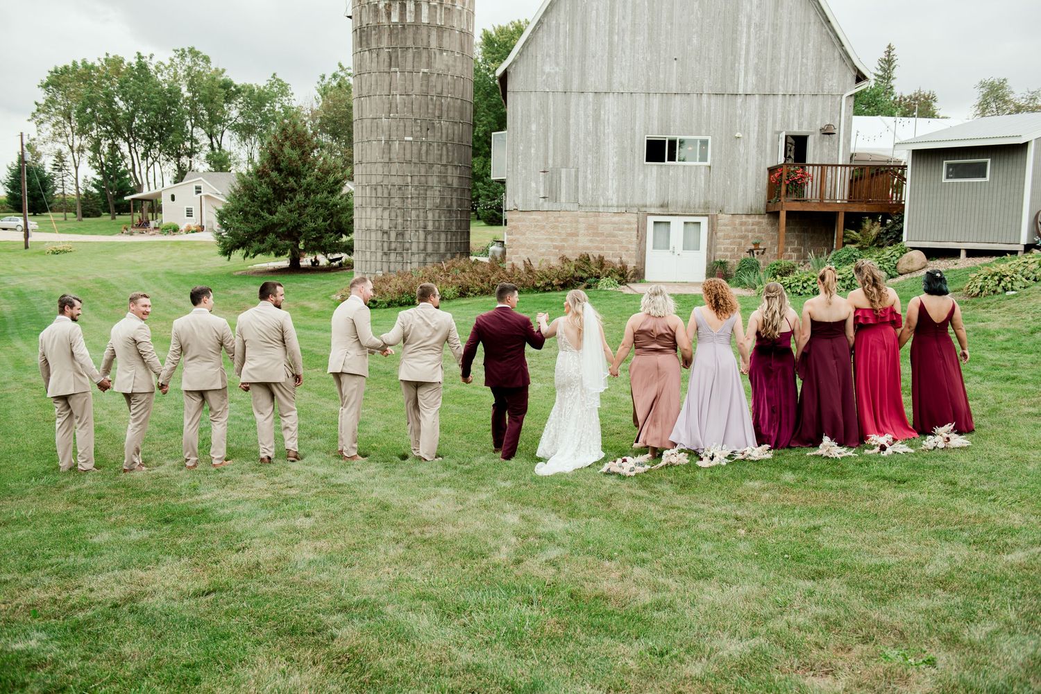 Wedding party lines up near rustic barn for formal group photo in burgundy and neutral tones.