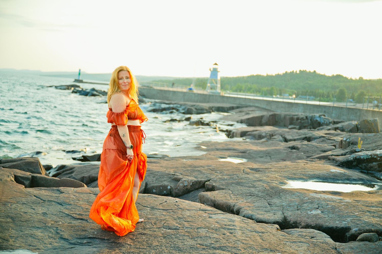 Woman in flowing orange dress standing on rocky shoreline with lighthouse and calm sea in background.