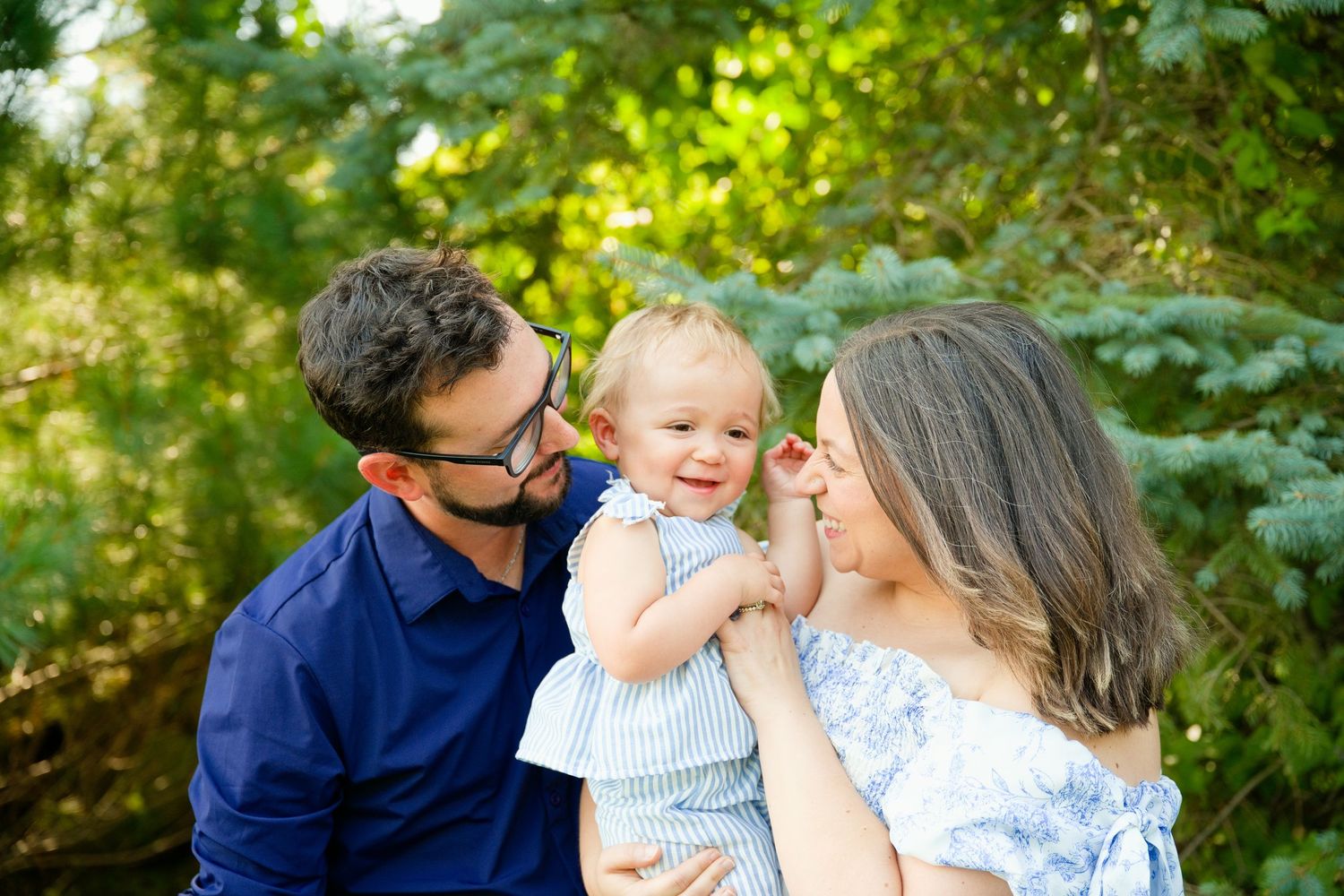 A happy family with a smiling baby in a blue outfit, posing outdoors among lush green trees.