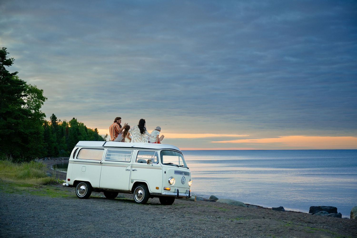 Friends sitting atop a vintage white VW van on a rocky beach at sunset, overlooking a calm ocean horizon.