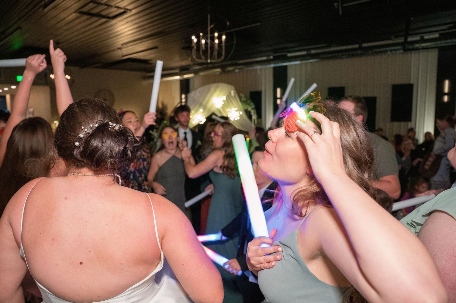 Wedding reception party with guests dancing and holding glow sticks under dim lighting.