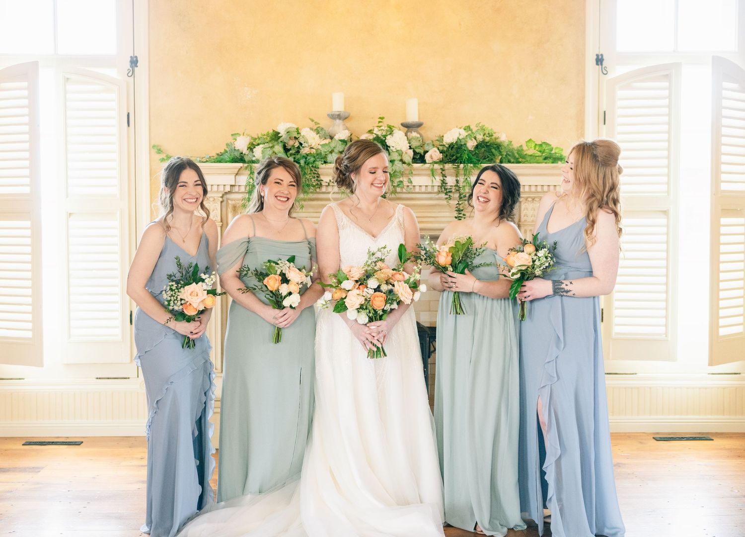Smiling bride with four bridesmaids in dusty blue gowns holding colorful floral bouquets indoors.