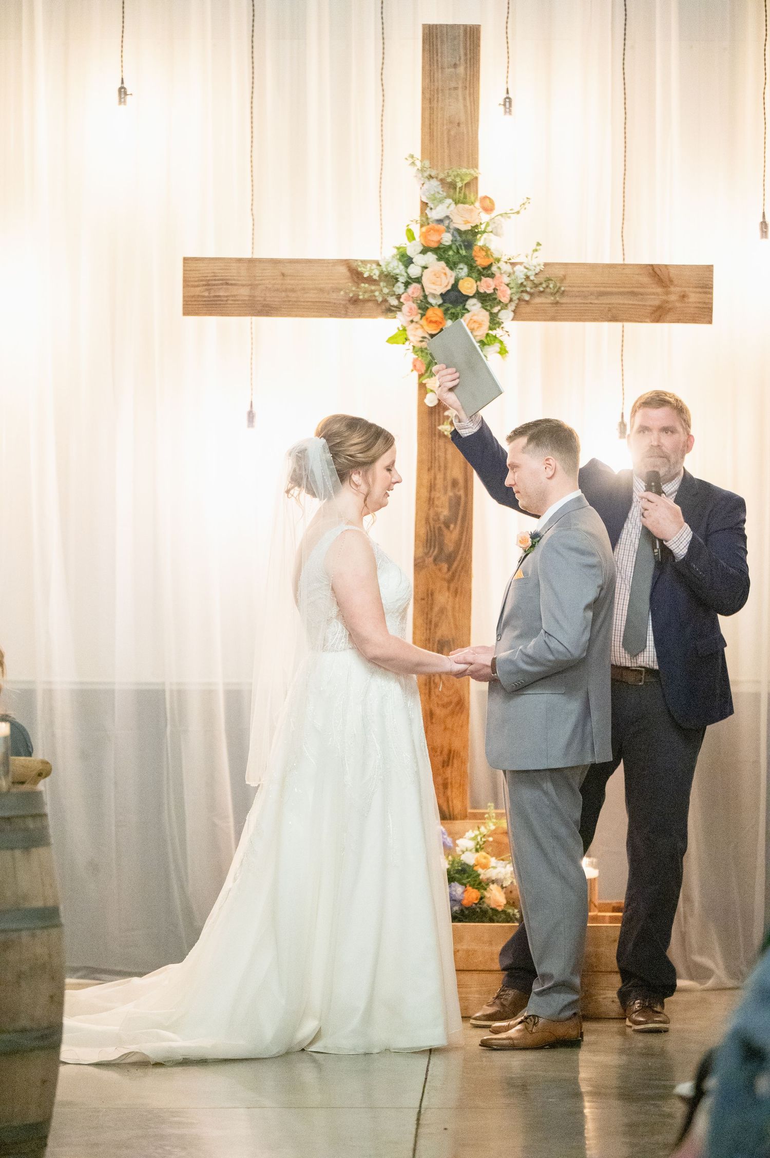 Groom raising Bible as officiant leads vows before wooden cross with floral arrangement.