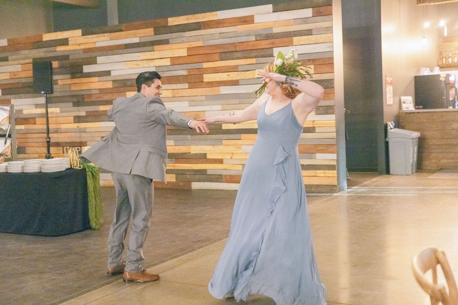 Bridesmaid in blue gown dances with groomsman in gray suit at wedding reception.