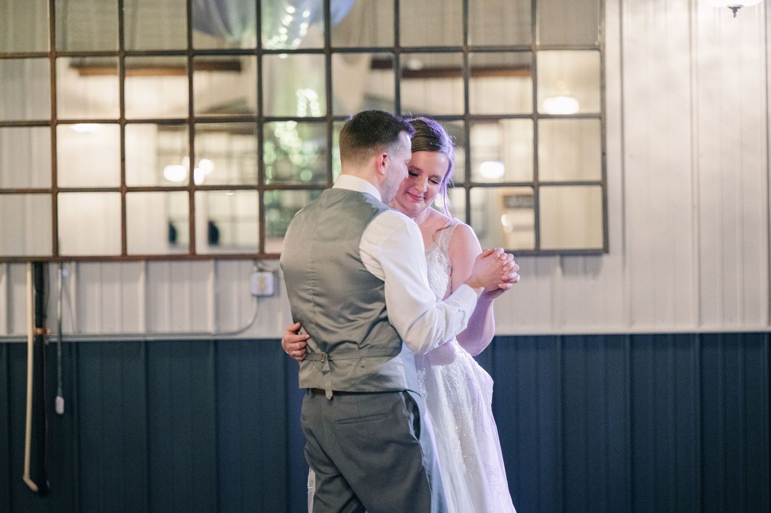 Bride and groom sharing intimate first dance embrace in industrial venue with large windows.