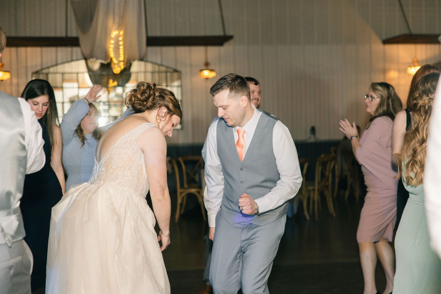 Bride in white gown and groom in gray vest dance energetically with guests on wedding reception dance floor.