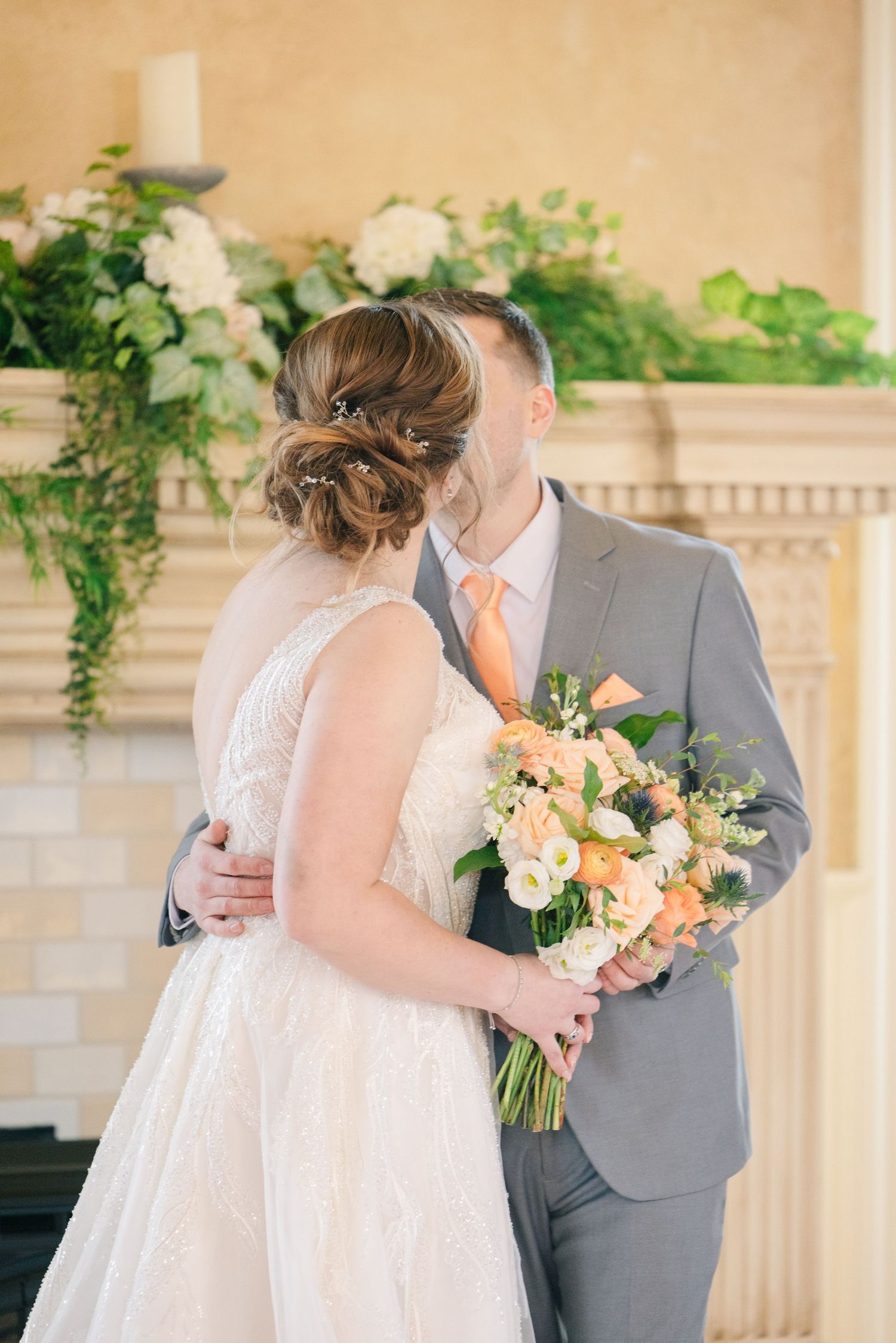 Bride and groom embrace during wedding ceremony, bride holding peach floral bouquet, groom in gray suit with orange tie.