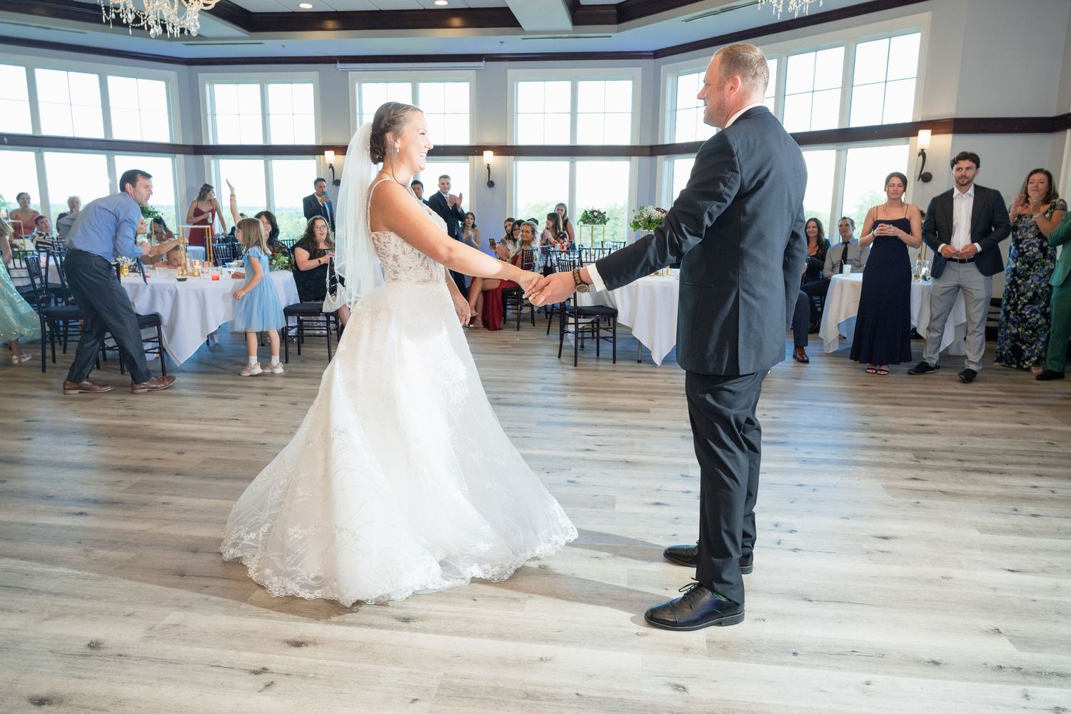 Bride and groom share their first dance on a hardwood floor surrounded by seated wedding reception guests.