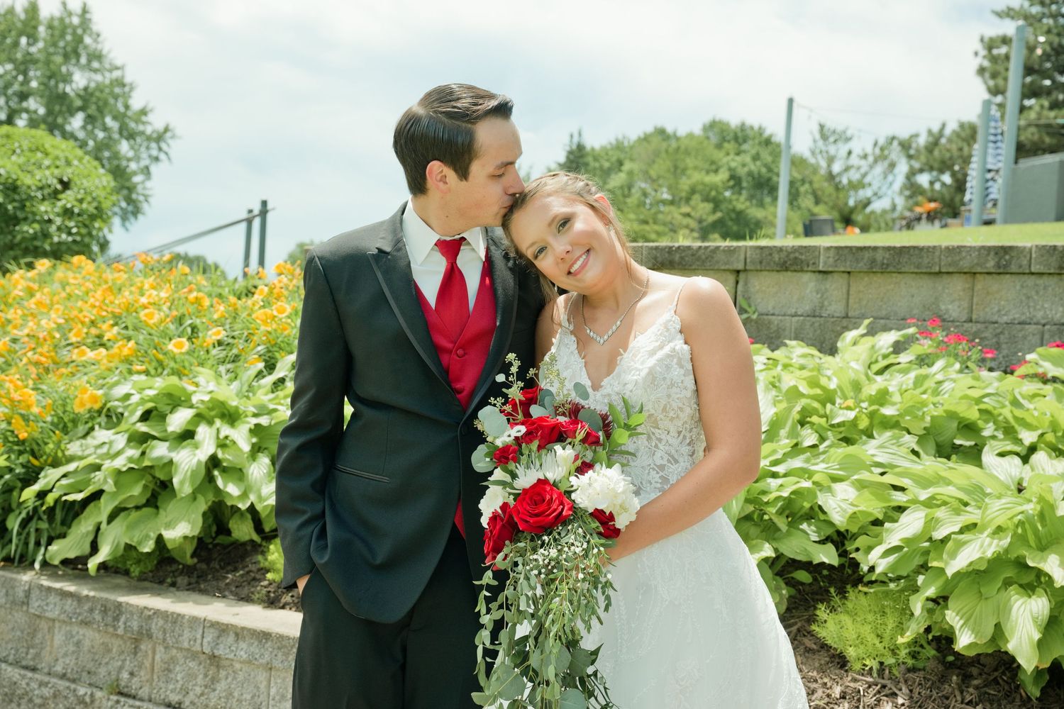 Bride and groom smiling together outdoors, holding red and white bouquet, surrounded by lush garden greenery.