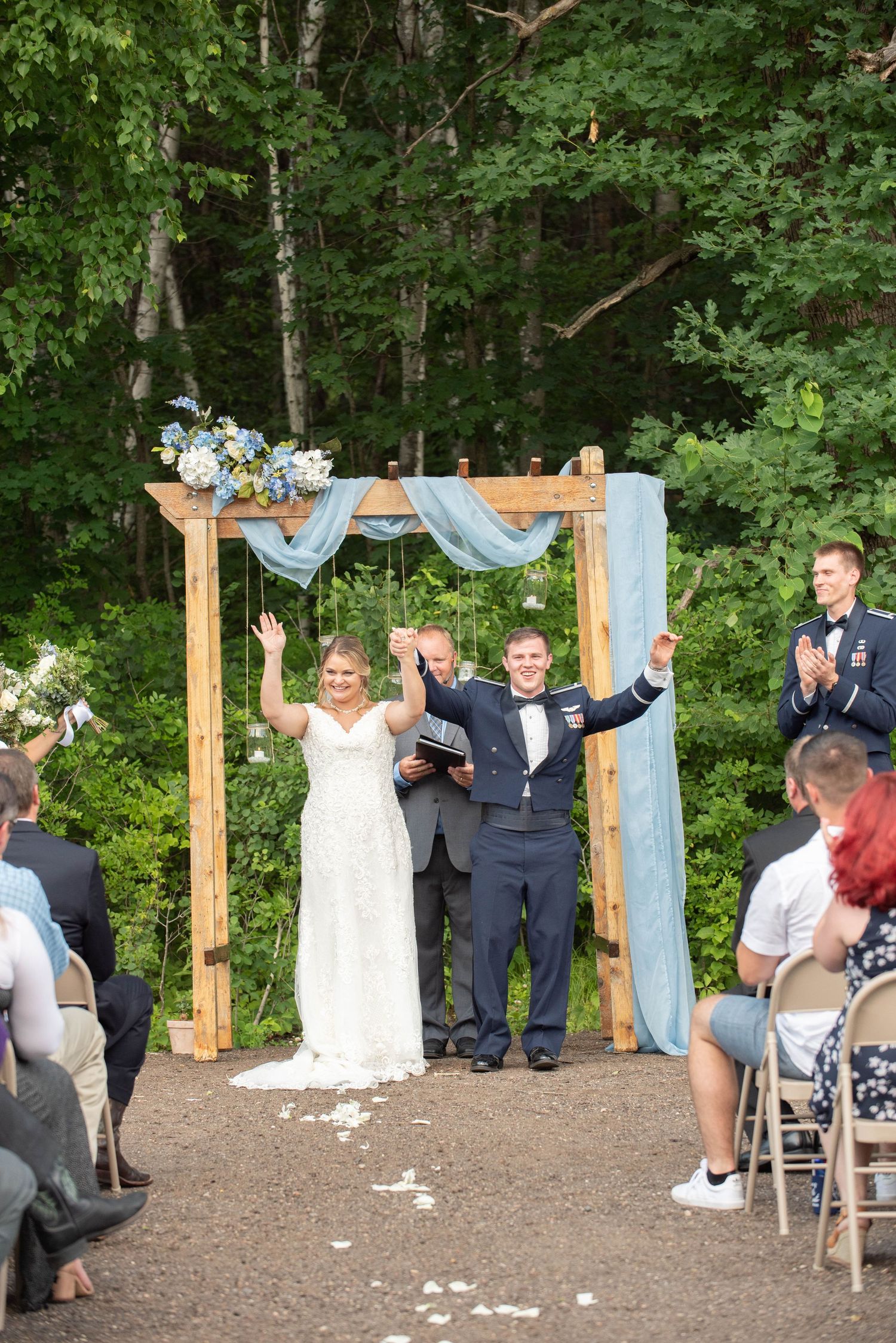 A wedding ceremony takes place under a wooden arch decorated with blue fabric and flowers in a wooded outdoor setting. Zimmerman MN Weddings