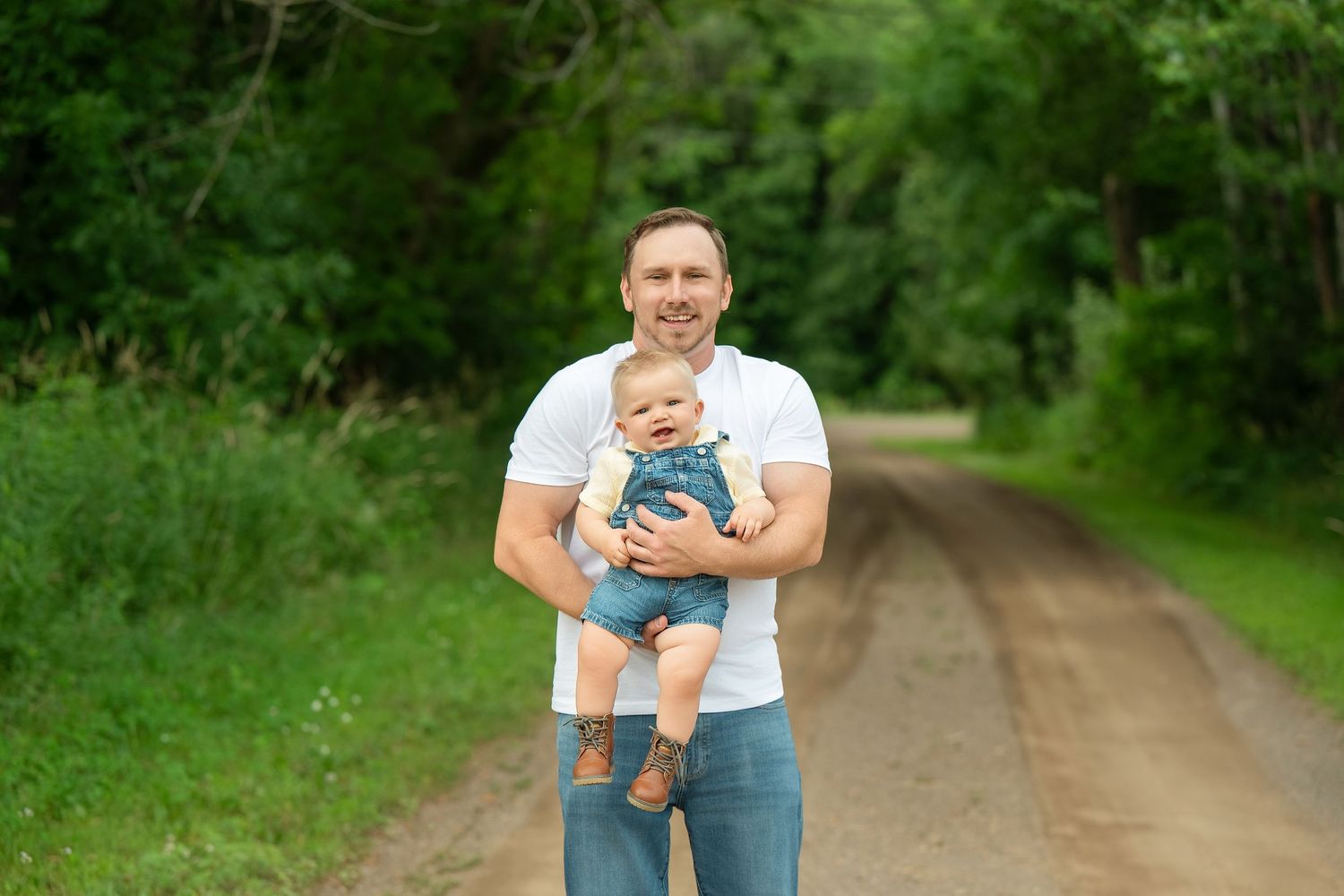 A dirt path through green foliage with an adult holding a small child in casual summer attire. Family Portraits & Milestone Moments in Fores