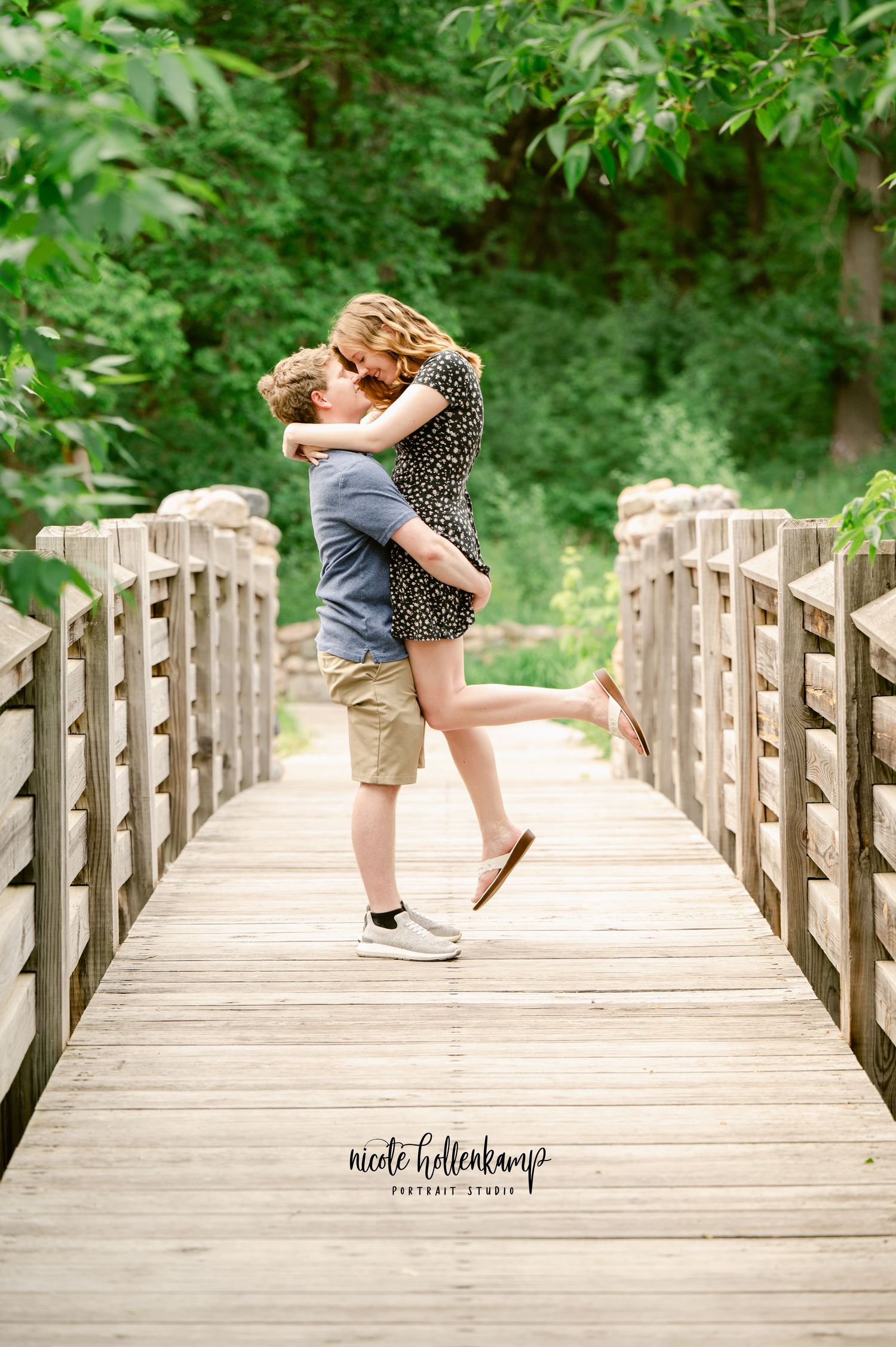 A romantic couple embraces and spins on a wooden bridge surrounded by lush green trees on a summer day.