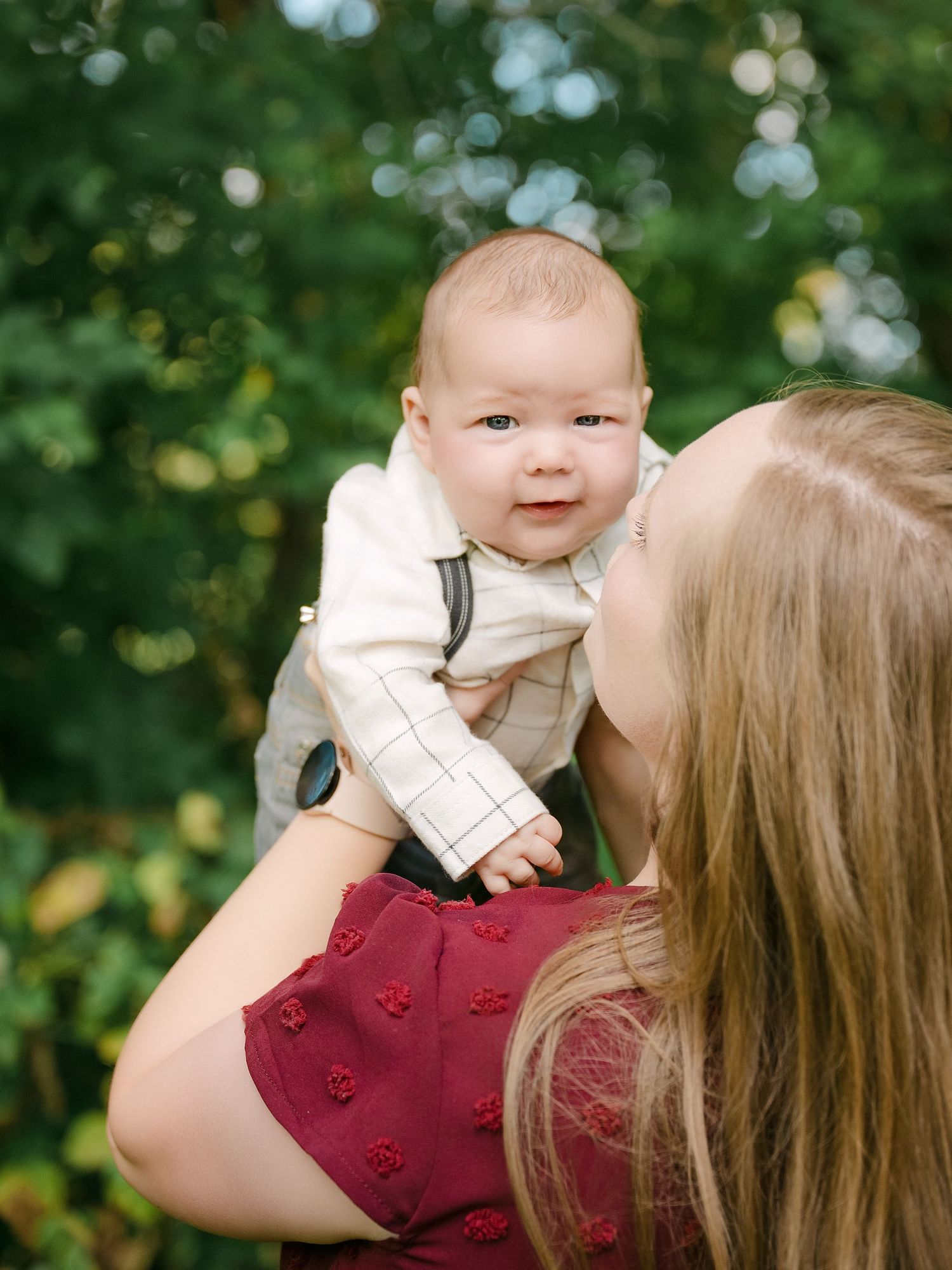 A mother lifts her smiling baby boy outdoors surrounded by lush green foliage.. Minnesota milestone photographer