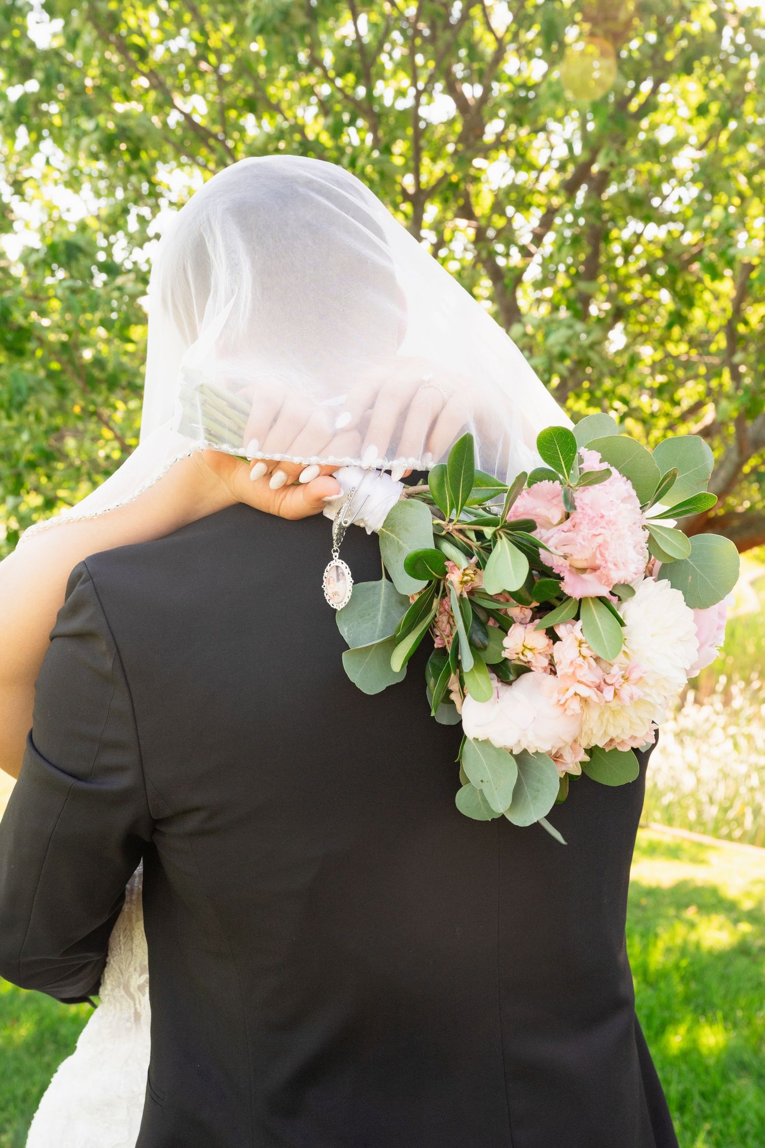 Bride embraces groom from behind holding pink and white bouquet with veil draped over both.