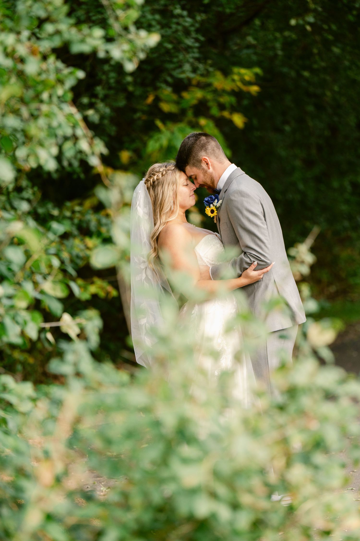 Central MN Wedding Photos | Bride and groom share romantic forehead touch surrounded by lush green foliage with veil flowing.