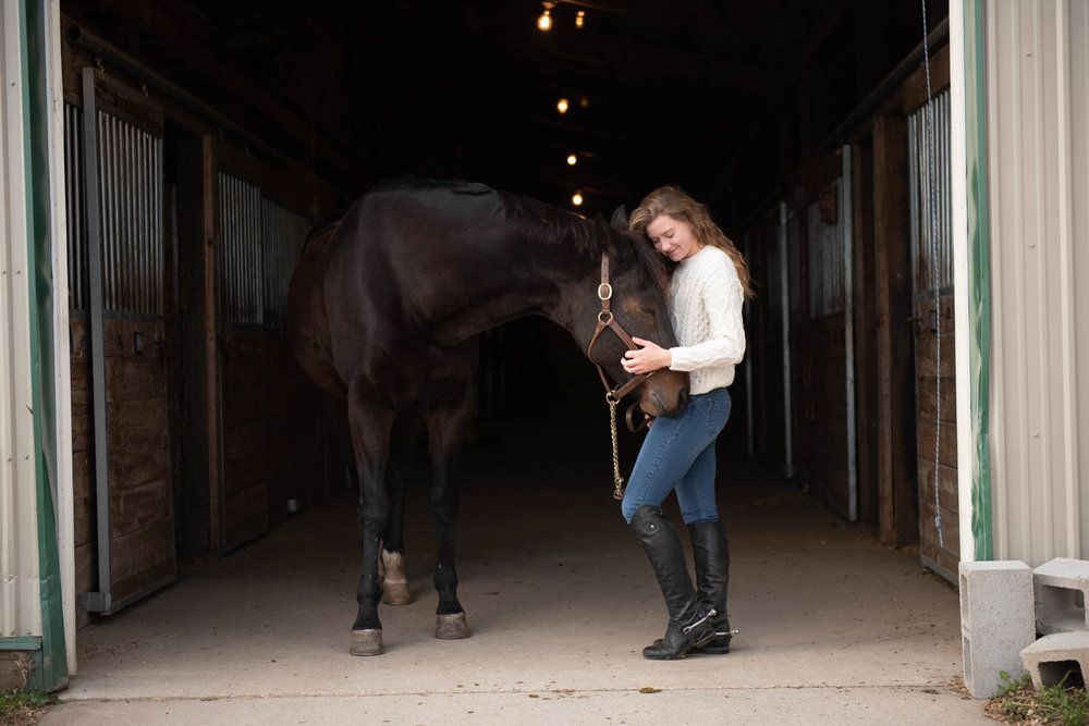 Central MN Senior Photography - Young woman in riding boots and jeans stands with a dark bay horse inside a stable barn aisle.