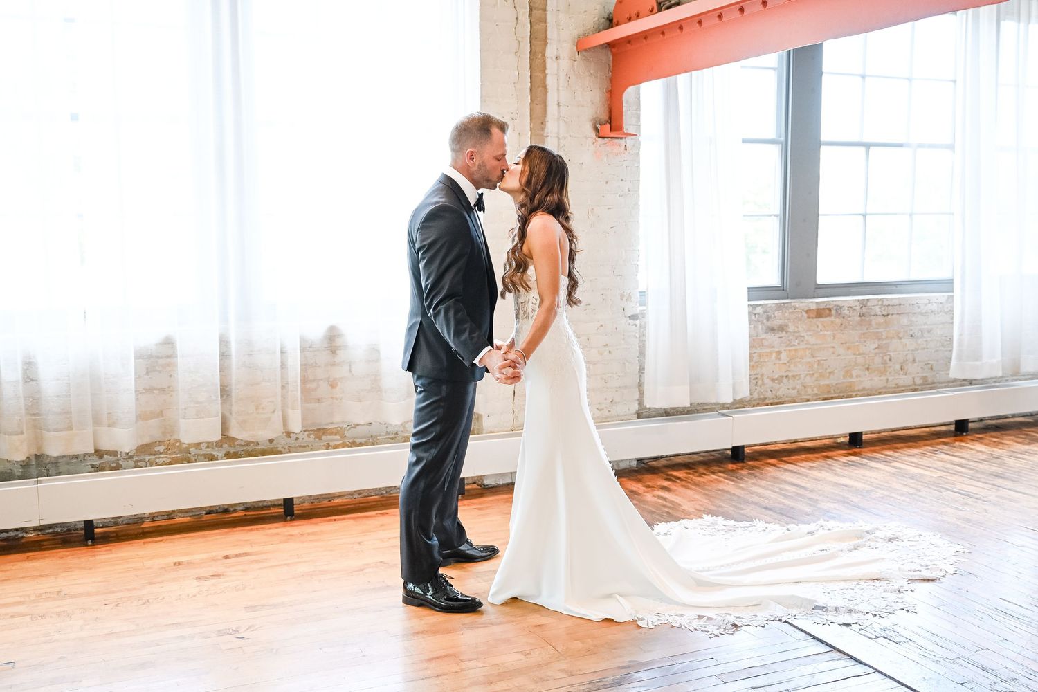 Central MN Wedding PhotBride and groom share a romantic first kiss in a sunlit industrial loft with hardwood floors and exposed brick walls.
