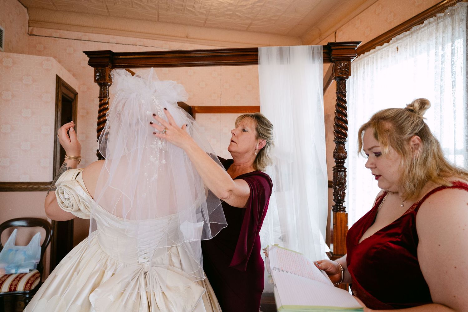 Central MN Wedding Photos | Two bridesmaids in burgundy velvet dresses help a bride arrange her veil before the wedding ceremony.