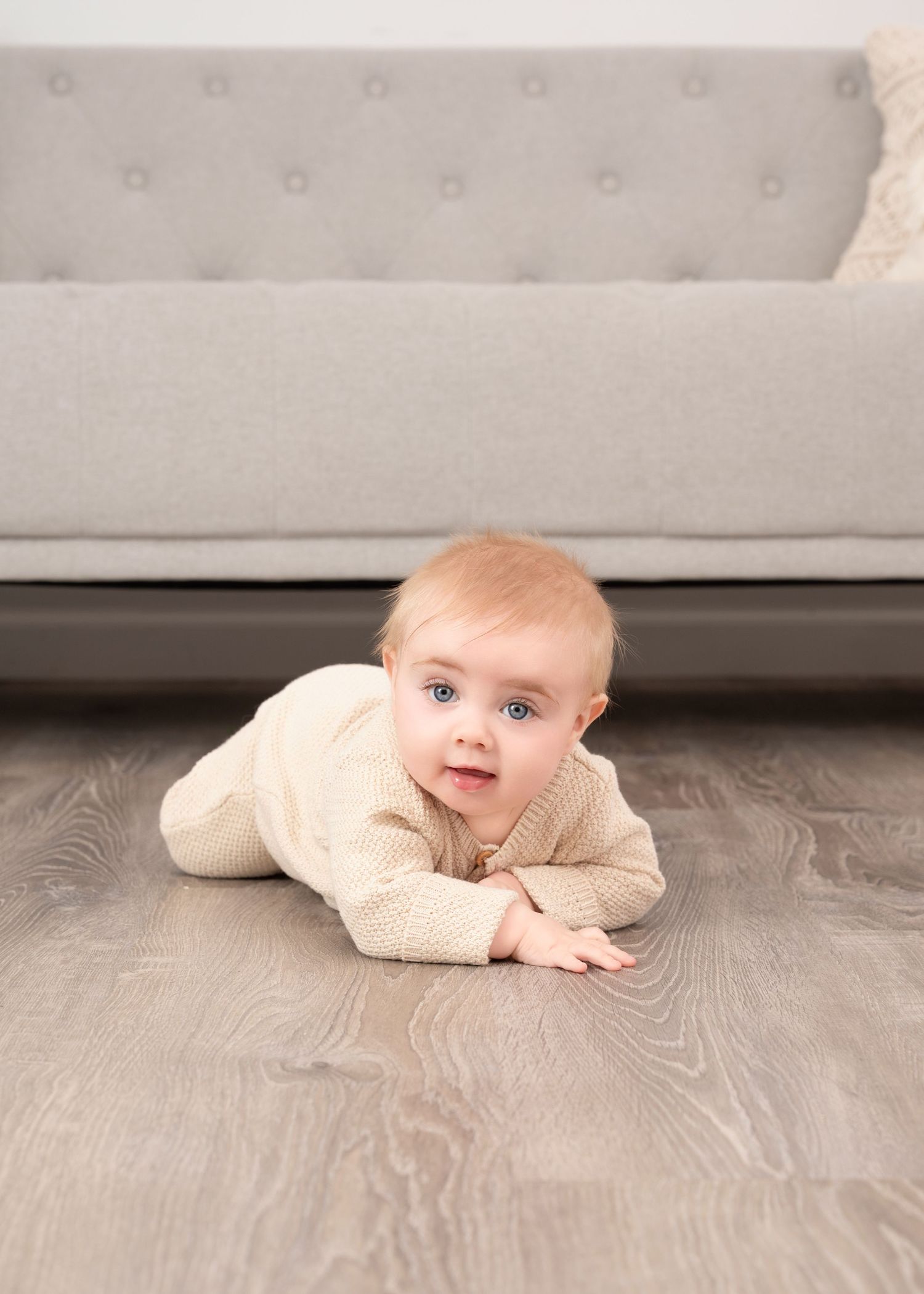 Central MN Milestone Photography -A cute baby with blue eyes in a cream knit outfit lies on a gray hardwood floor in front of a modern sofa.