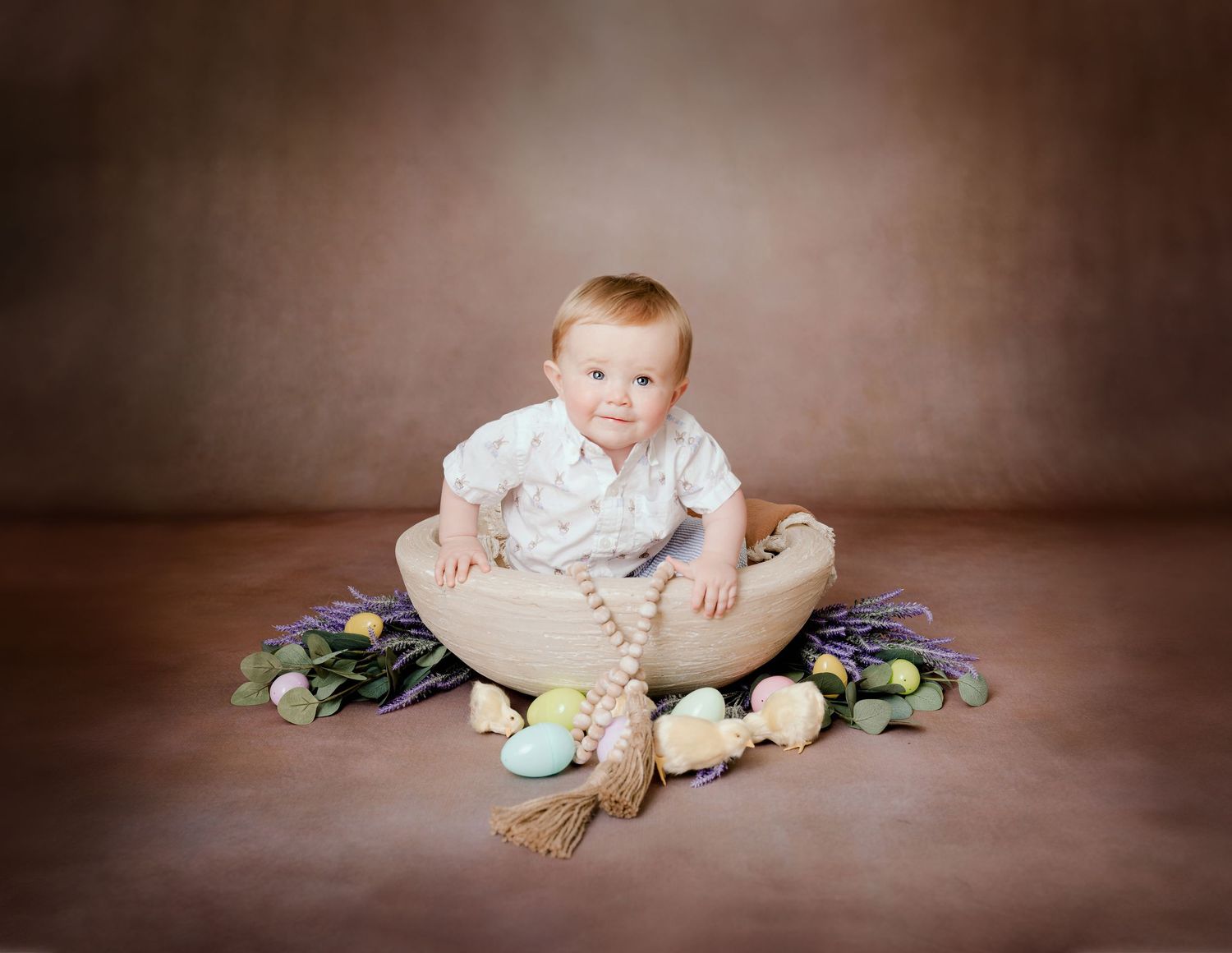Central MN Milestone Photos - Baby boy in white outfit sits in wooden bowl surrounded by Easter eggs and spring flowers on brown backdrop.