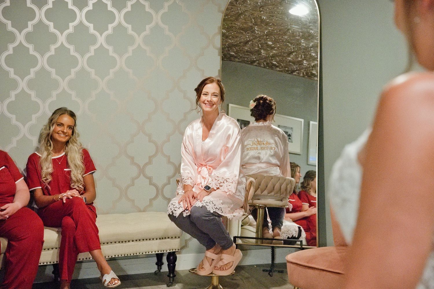 Smiling woman in pink satin robe sits on bench reacting to bride trying on wedding dress.