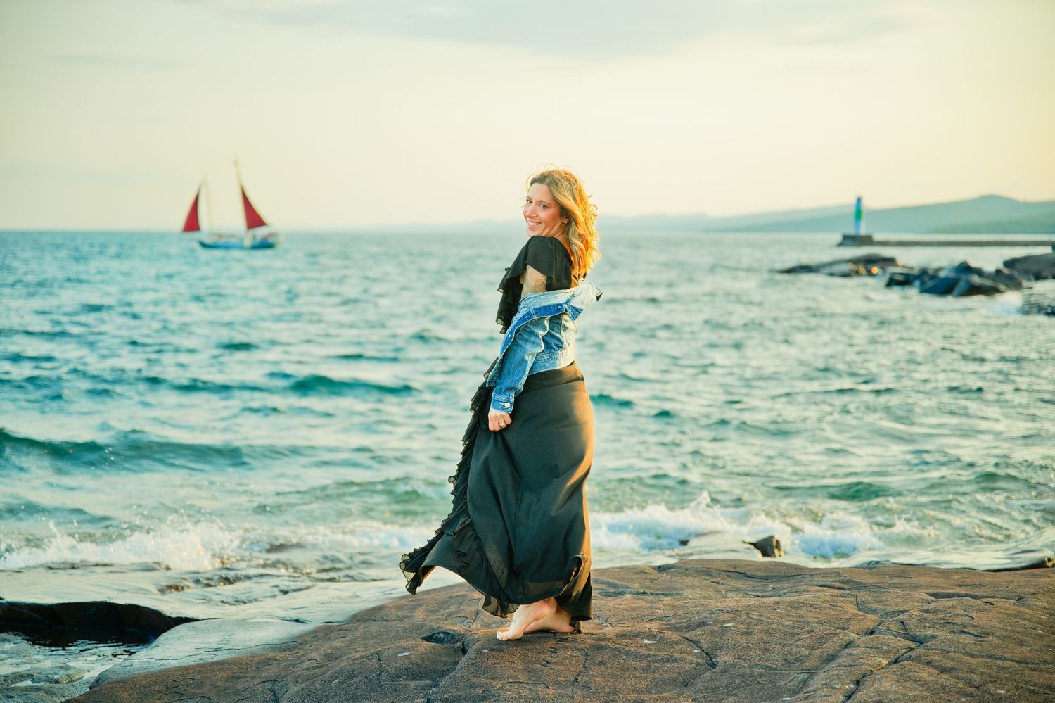 Woman in a long olive dress and denim jacket stands barefoot on a rocky shore at sunset with red-sailed boats on the sea.