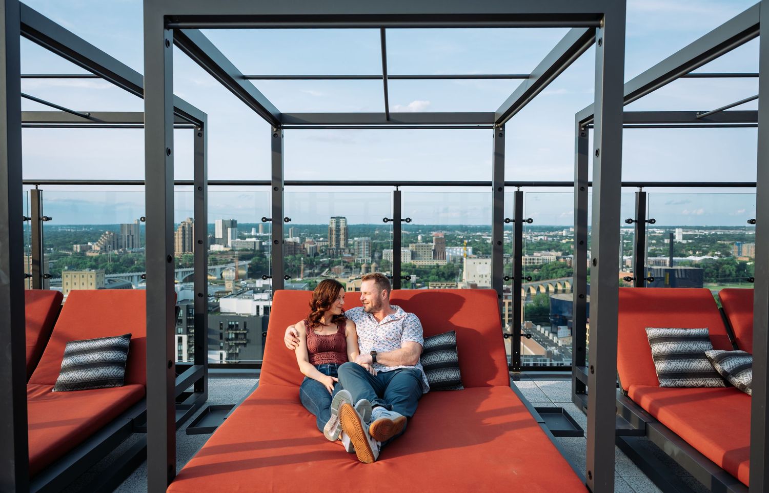 A couple relaxes on red cushioned seating with panoramic city views through glass windows.