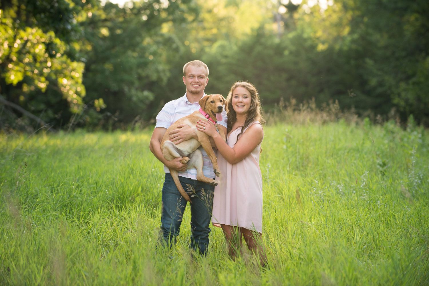 A couple stands together in a sunlit meadow holding their golden retriever puppy during a summer evening photo session.