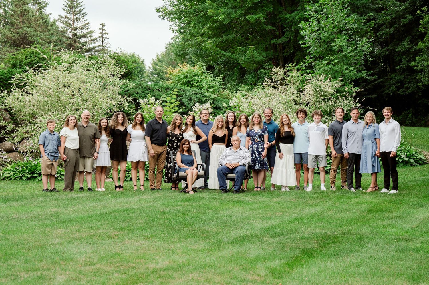 Large group wearing coordinated outfits poses together on green lawn with flowering bushes and trees in background.
