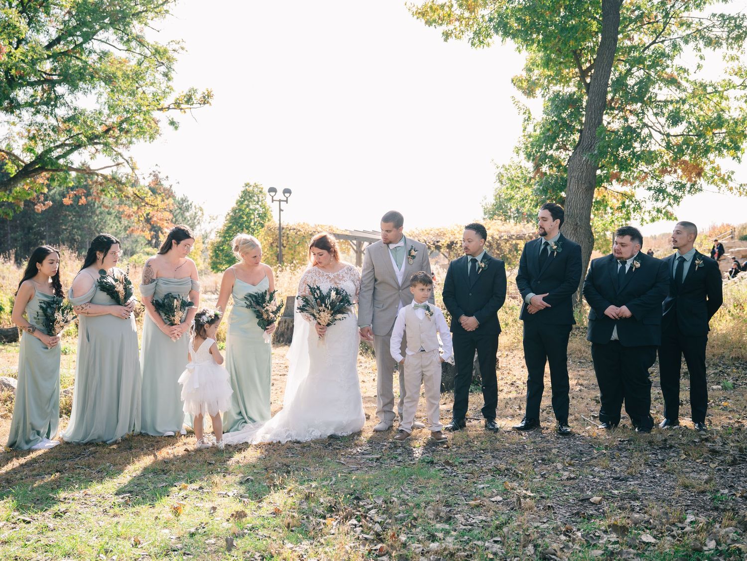 Wedding party poses outdoors in fall foliage, bridesmaids in sage green gowns and groomsmen in black suits flanking the bride and groom.