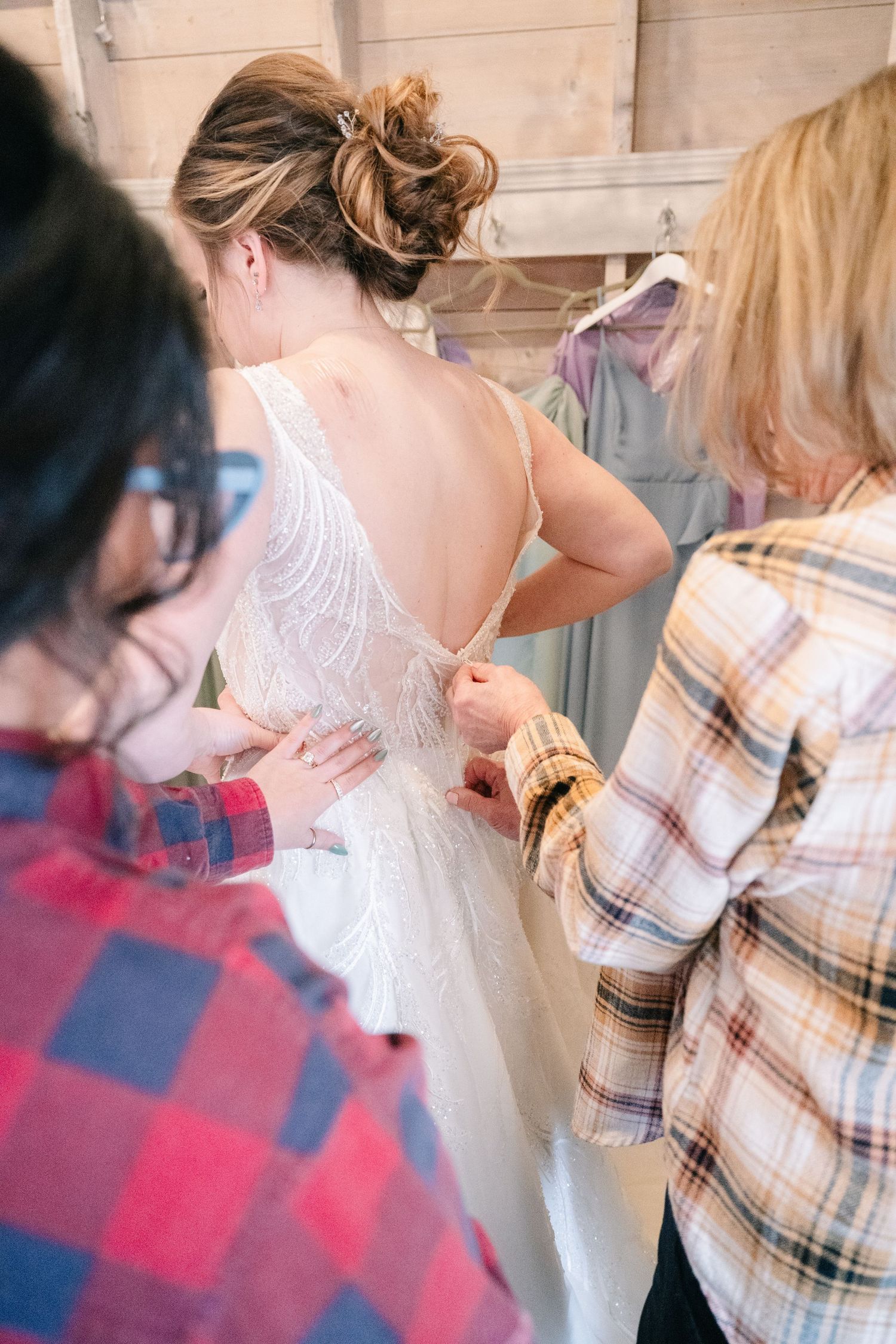 Two women helping a bride fasten her open-back lace wedding dress in a rustic bridal suite.