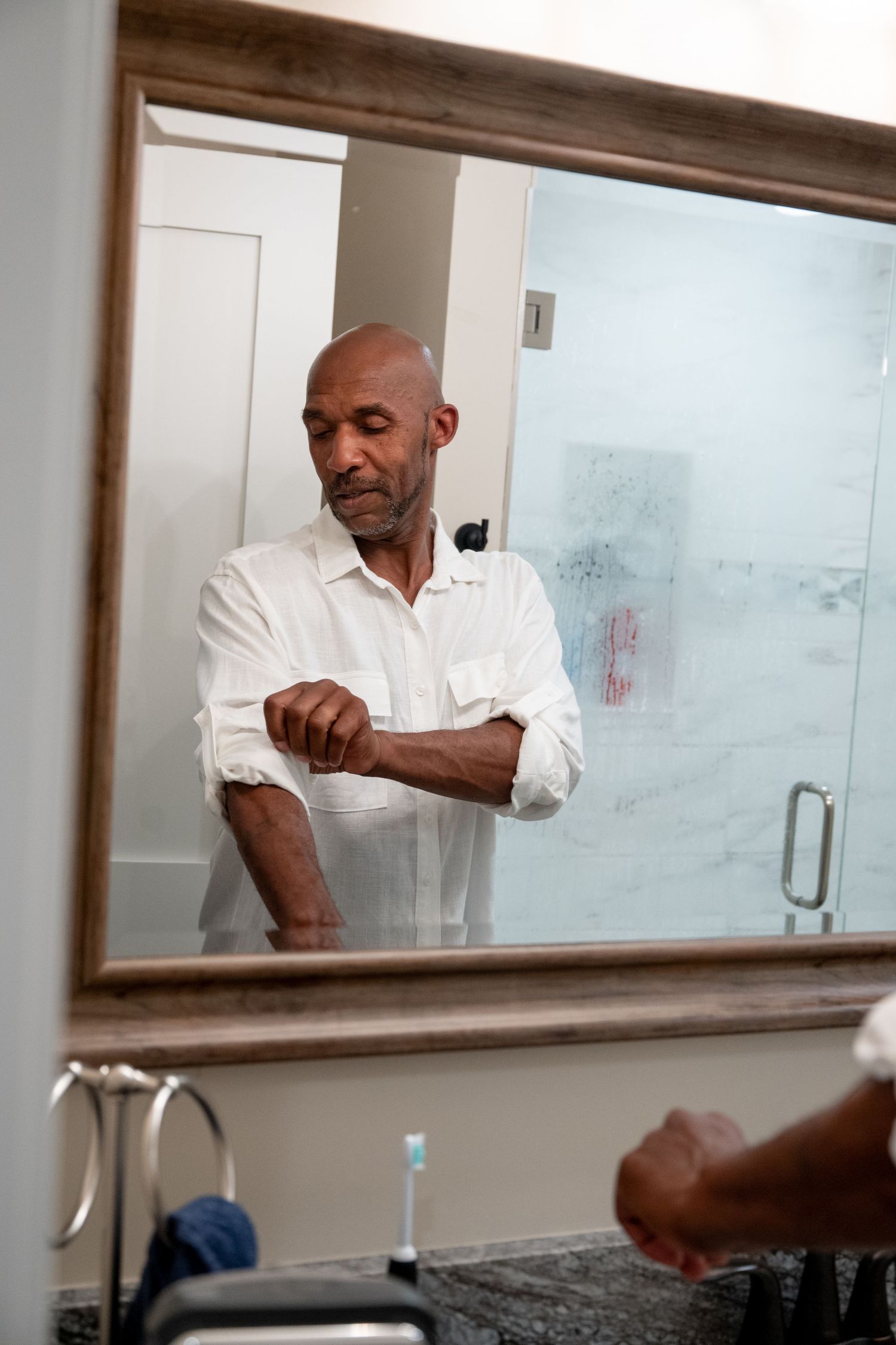 Bald man in white shirt rolling up his sleeve while looking in a bathroom mirror.