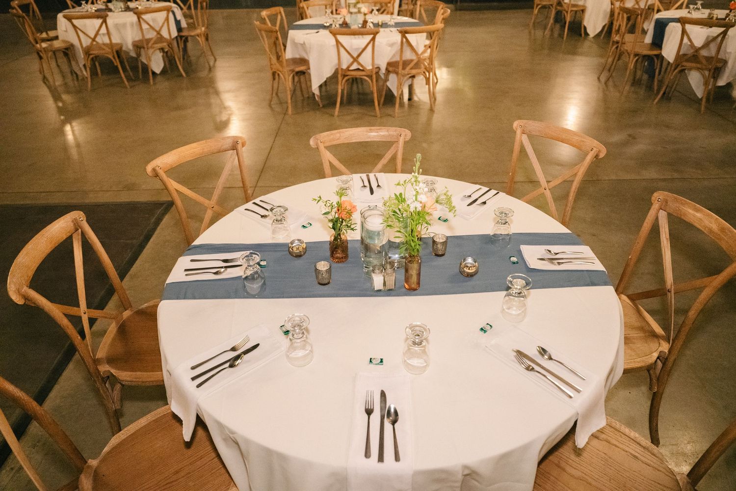 Elegant wedding reception tables with white linens, blue runners, wooden cross-back chairs, and floral centerpieces on concrete floors.