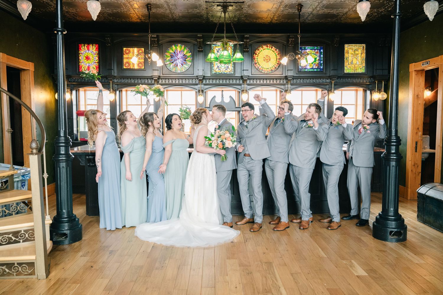 Wedding party in gray suits and blue dresses celebrate inside venue with stained glass windows.