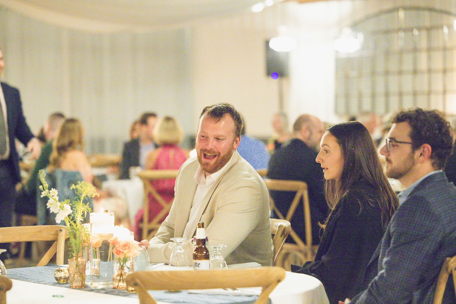 Wedding guests laughing and chatting at rustic wooden tables during reception dinner celebration.