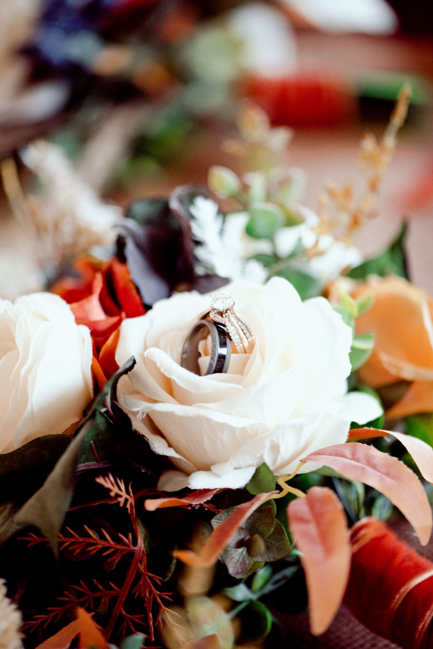 Wedding rings displayed atop a white rose in an elegant floral arrangement with red and peach accents.