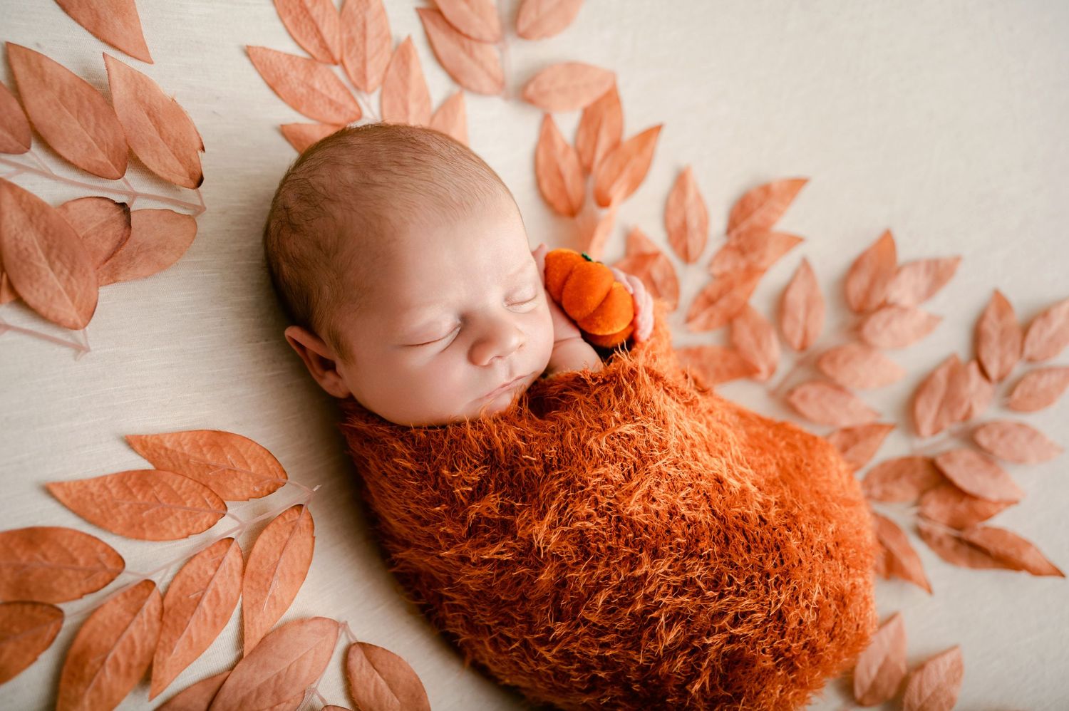 Sleeping baby wrapped in orange knit blanket surrounded by peach-colored autumn leaves in a dreamy portrait setting.