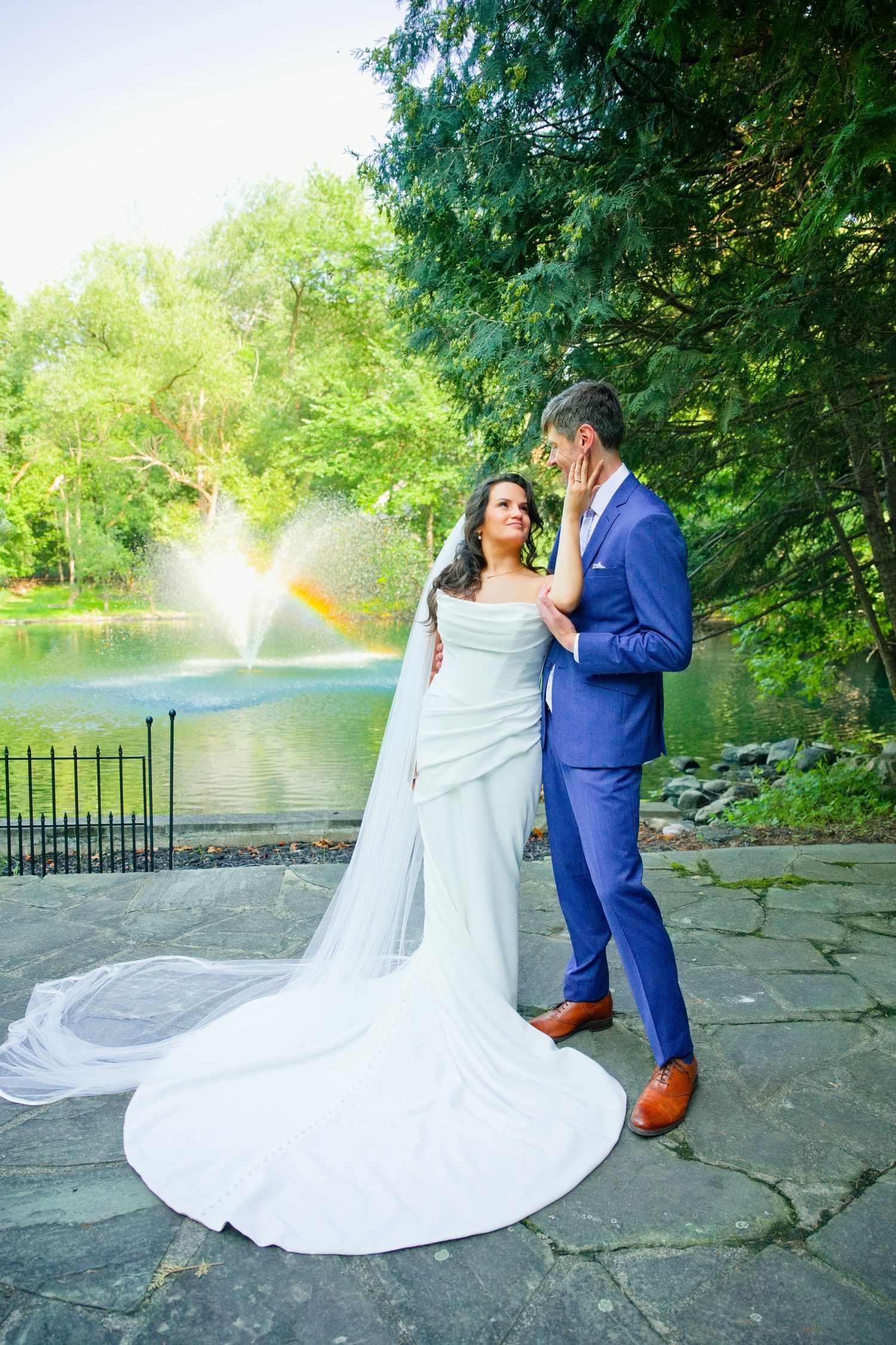 Bride in white gown with veil and groom in blue suit pose romantically by a fountain in a lush green park.