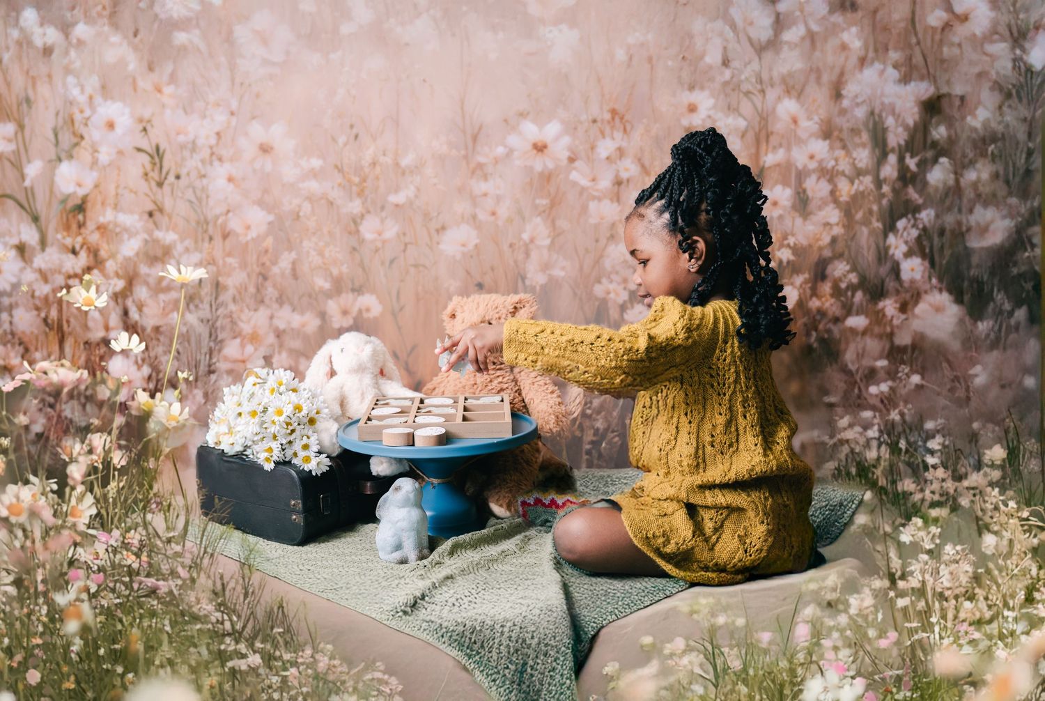 Young girl in yellow knit outfit reaches for macarons on blue cake stand among flowers with stuffed animals. mother day photoshoot