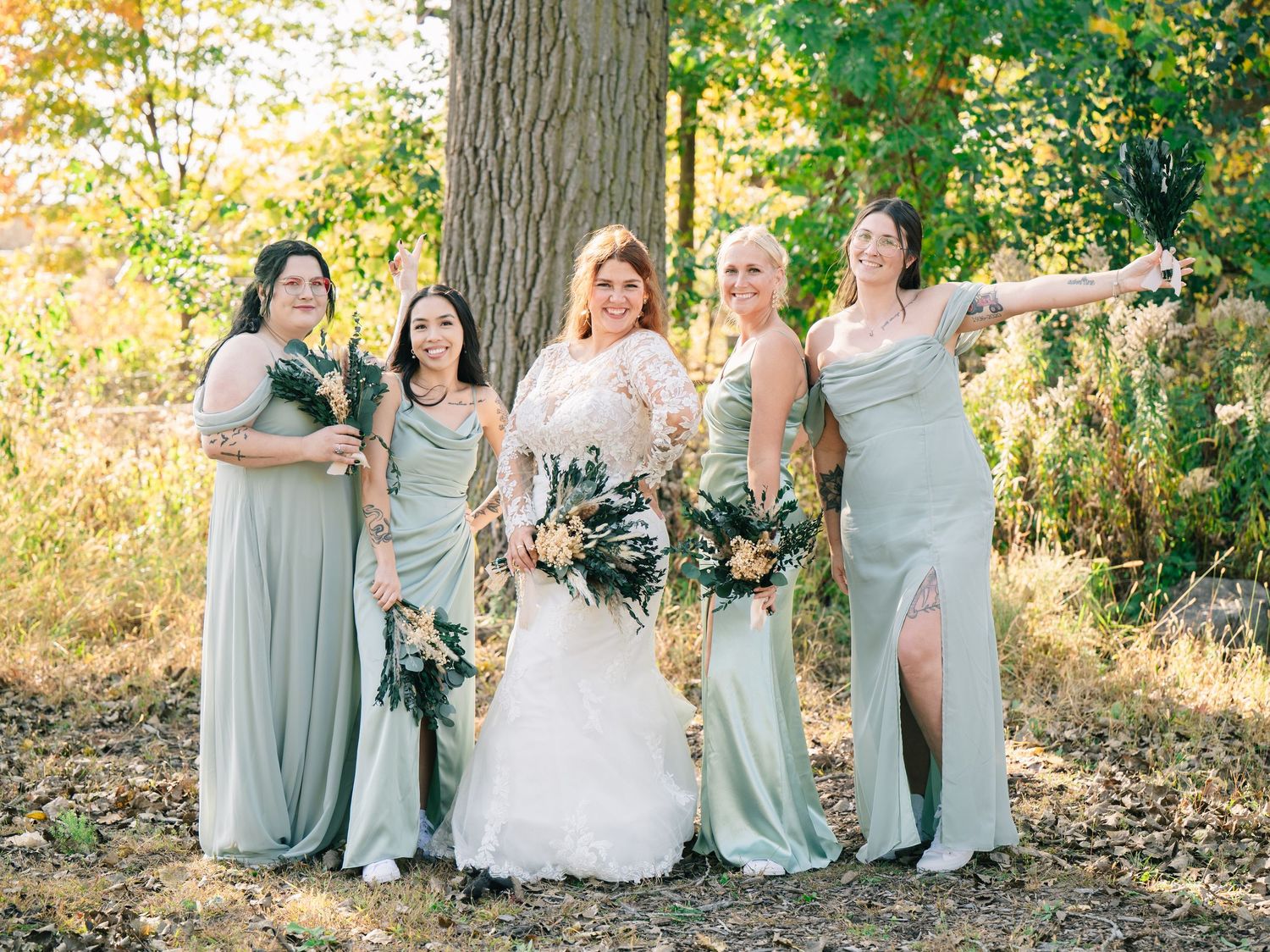 Bride in white lace gown poses with four bridesmaids in sage green dresses holding bouquets outdoors.