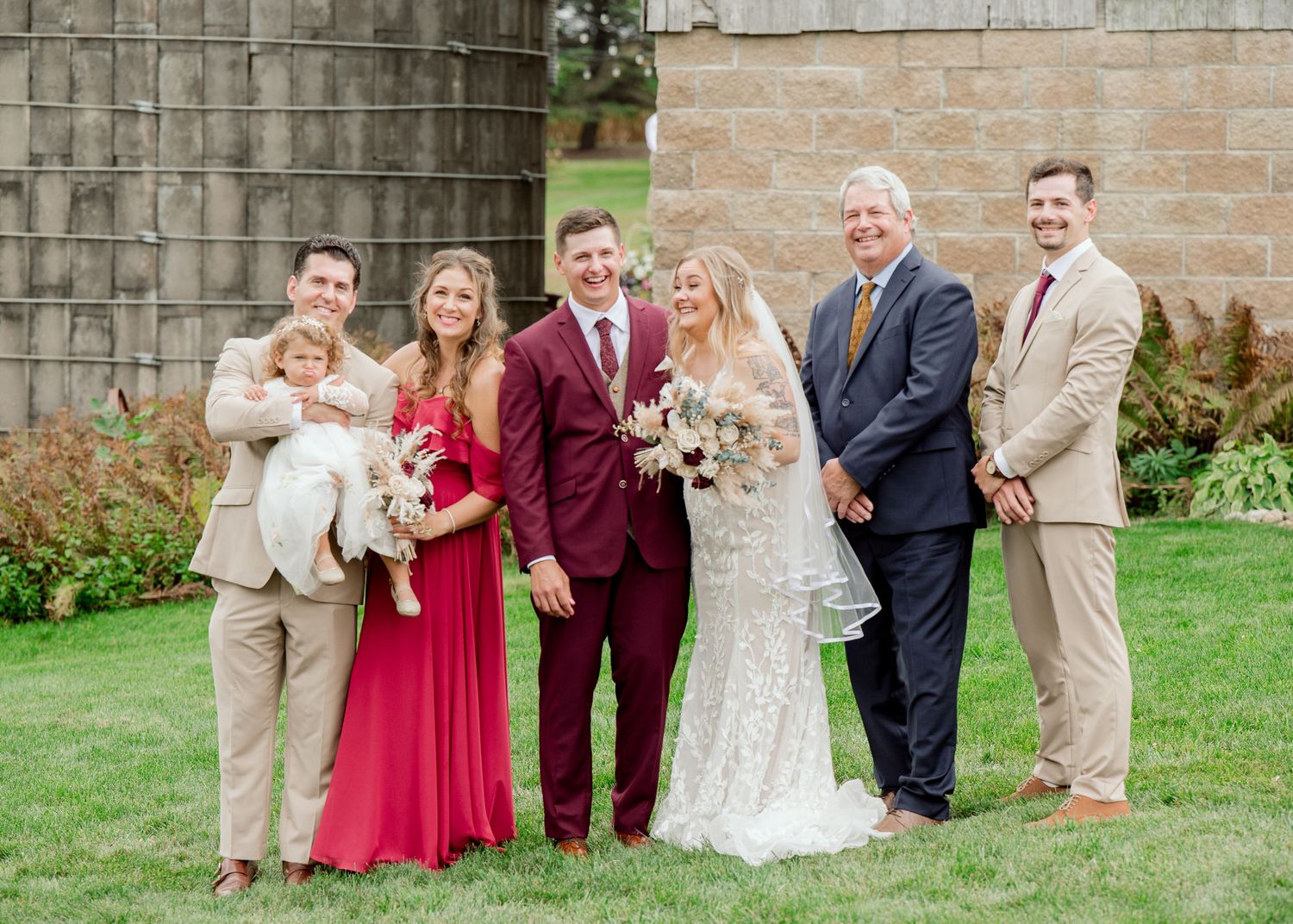 Wedding party in formal attire poses together on lawn near rustic stone wall with garden plants.