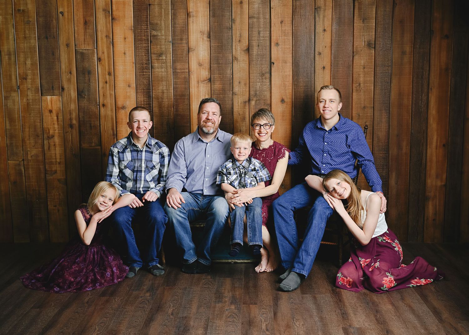 A family sits together against a rustic wood paneled wall wearing coordinated blue and burgundy outfits.