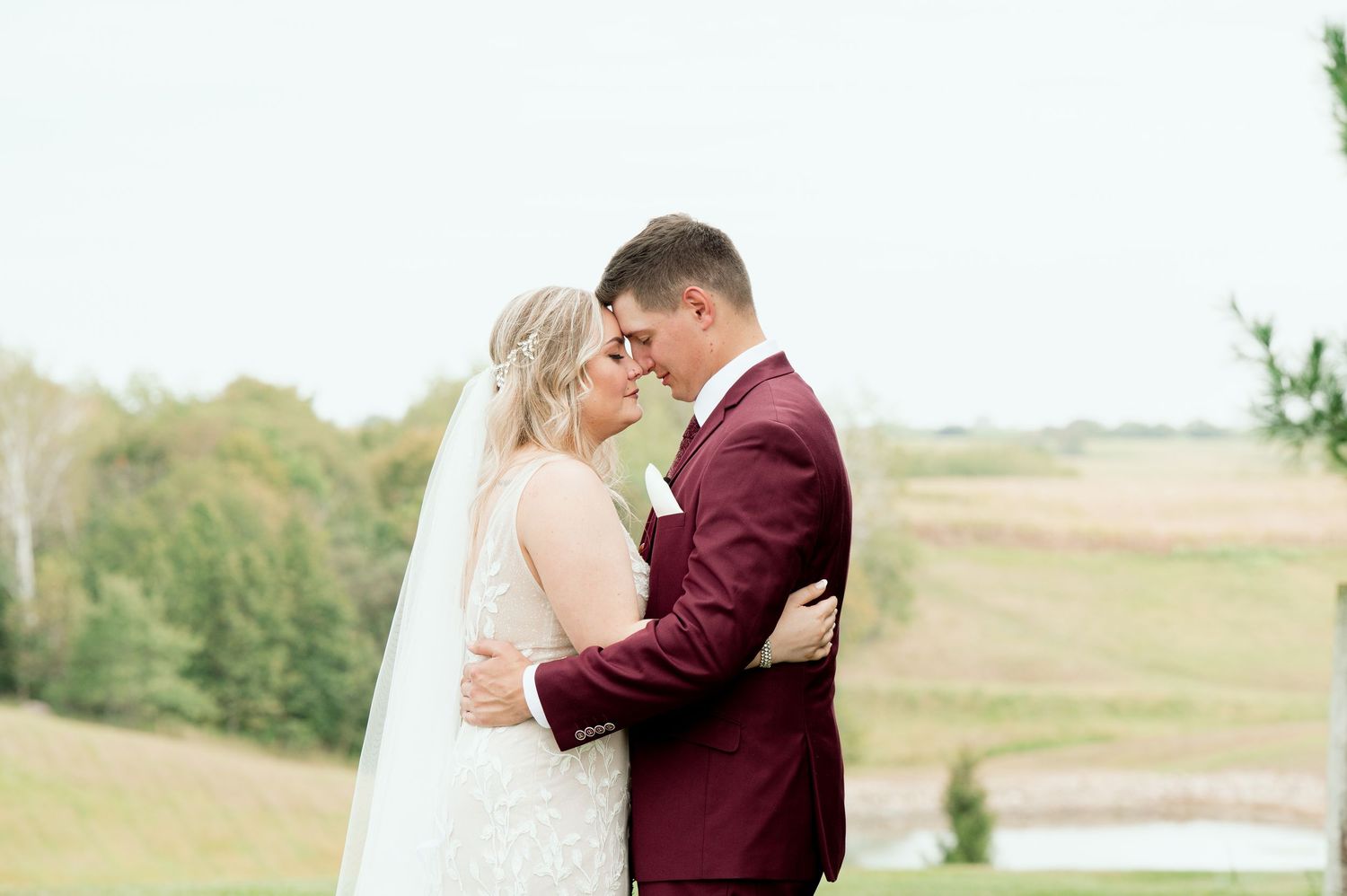 A romantic wedding couple embraces in a burgundy suit and white dress against a natural outdoor backdrop.