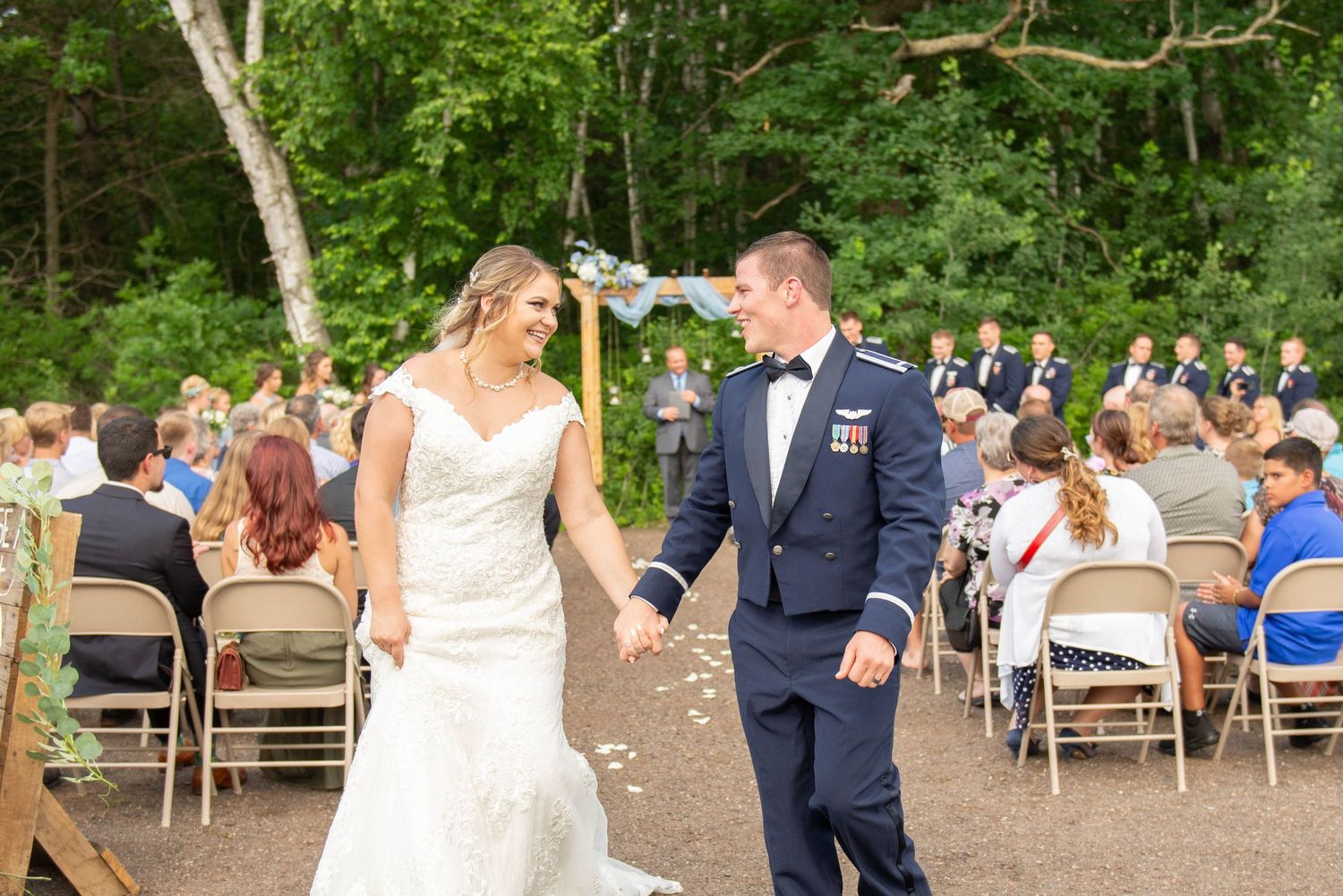 Outdoor wedding ceremony with guests seated on white chairs surrounded by lush greenery. Zimmerman MN Weddings