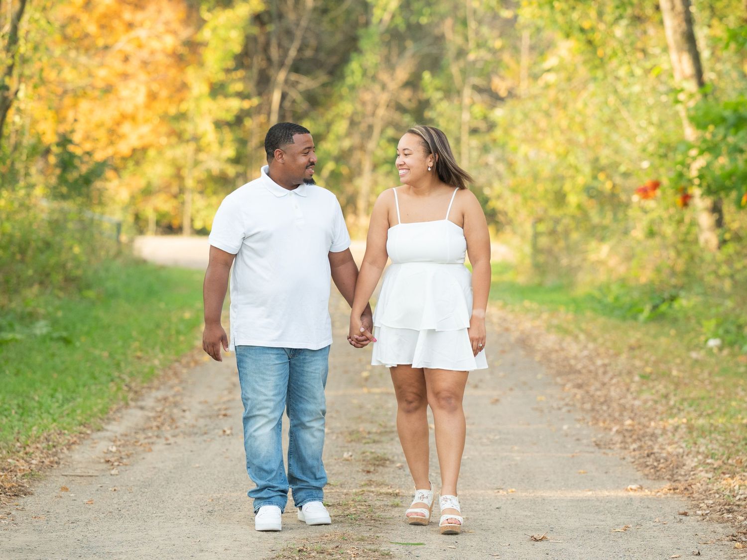 Central Minnesota Best Engagement Couple holding hands walking on a tree-lined path in autumn, wearing white outfits, smiling at each other.