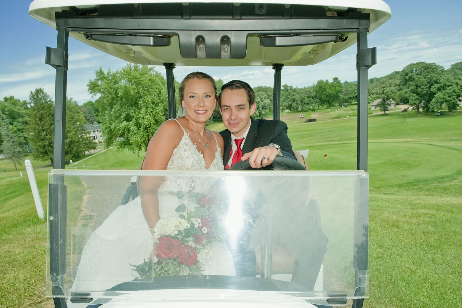 Bride and groom smile together while riding in a golf cart on a sunny green golf course.