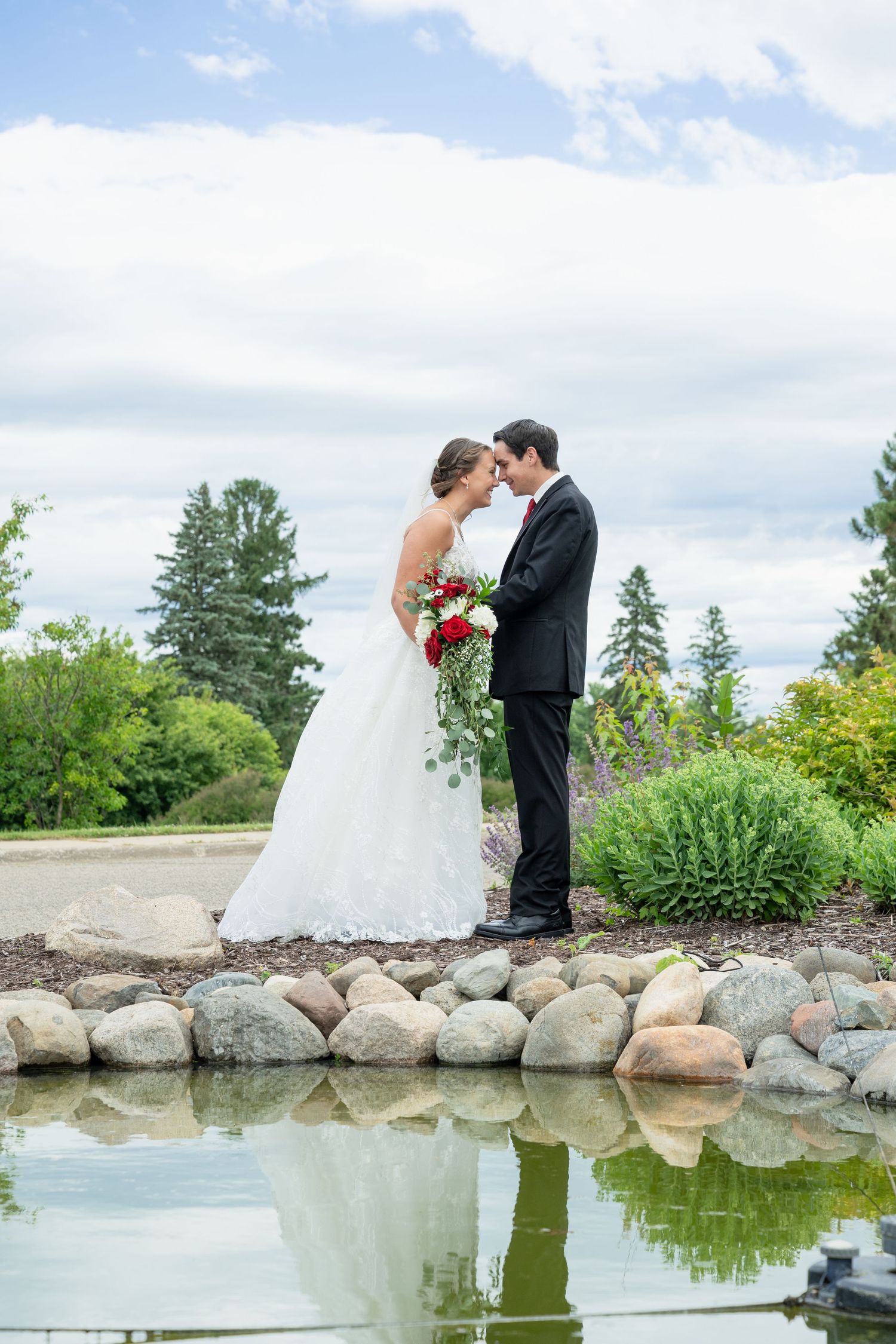 Bride and groom share a romantic moment by a reflective pond surrounded by lush greenery on their wedding day.
