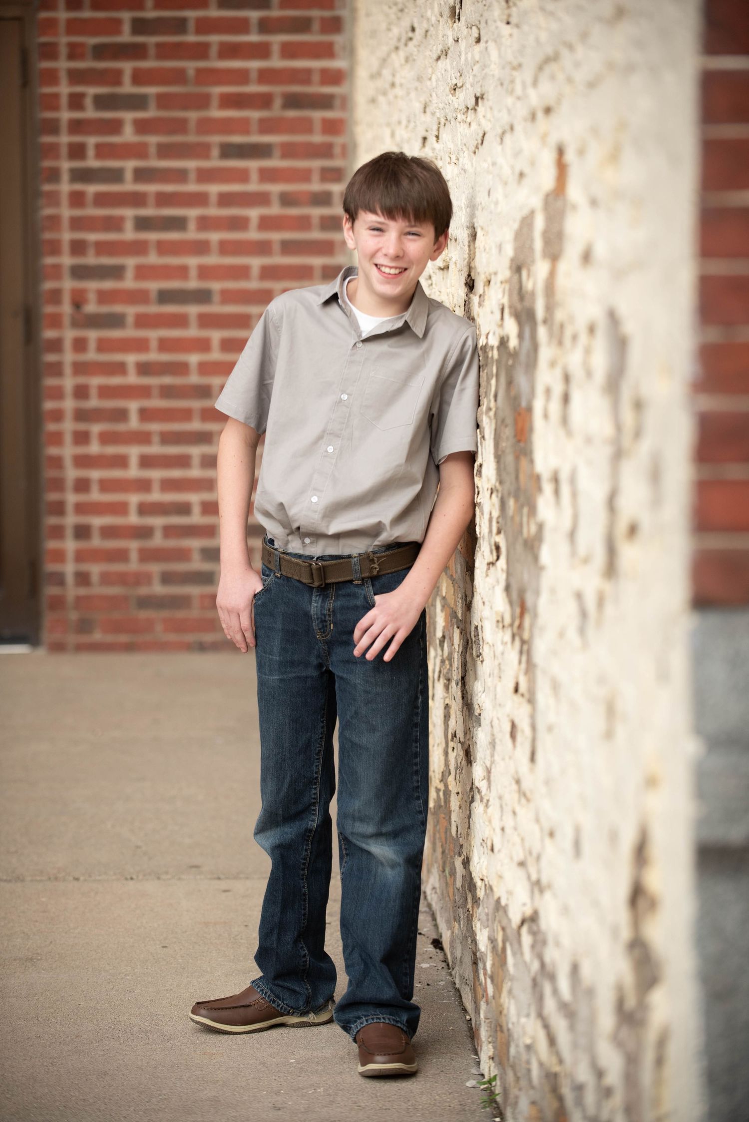 A young child in a gray shirt and jeans poses casually against a white pillar with a brick wall in the background.