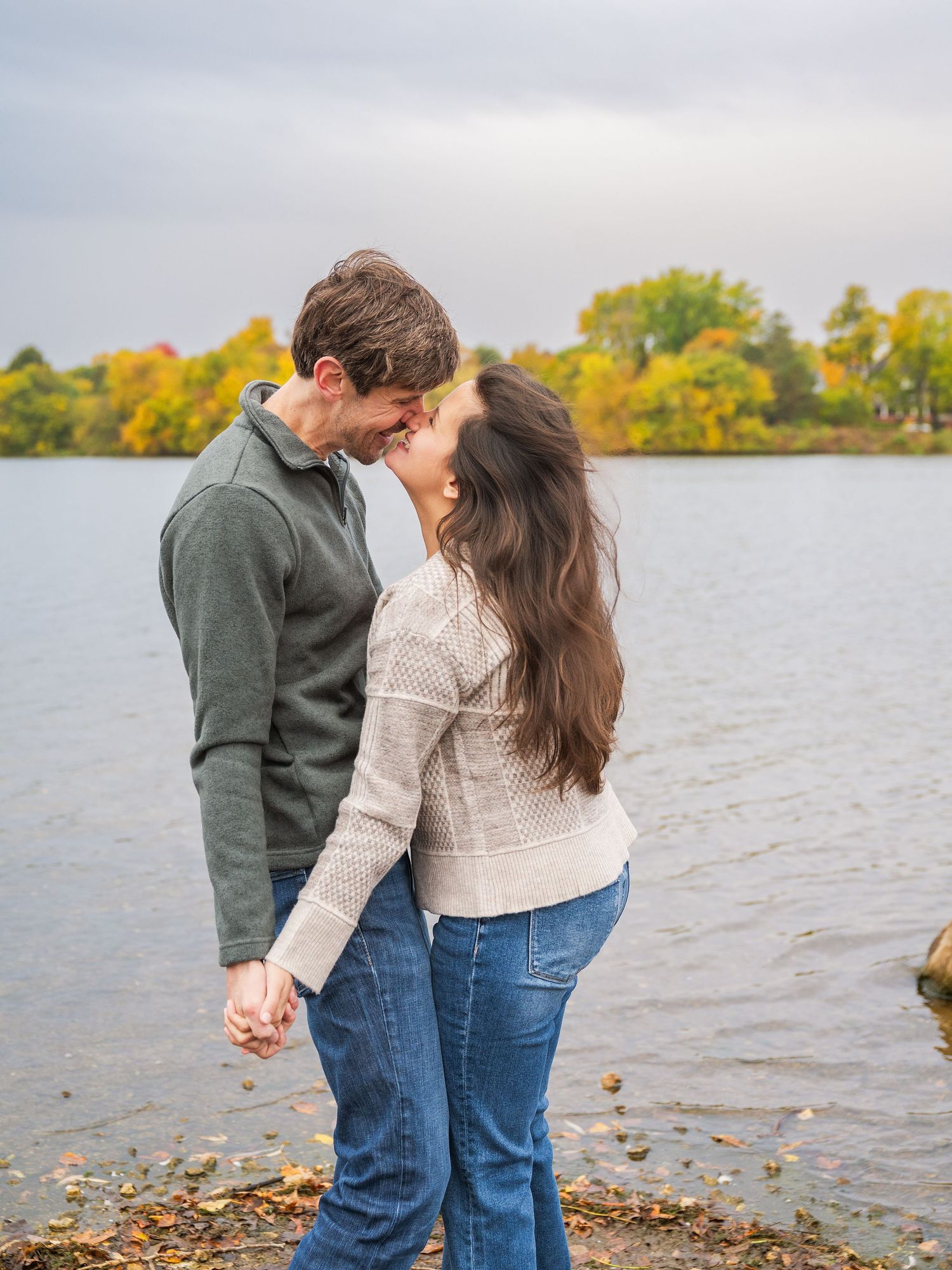 A couple in casual attire holds hands while walking along a scenic lakeside with autumn foliage in the background.